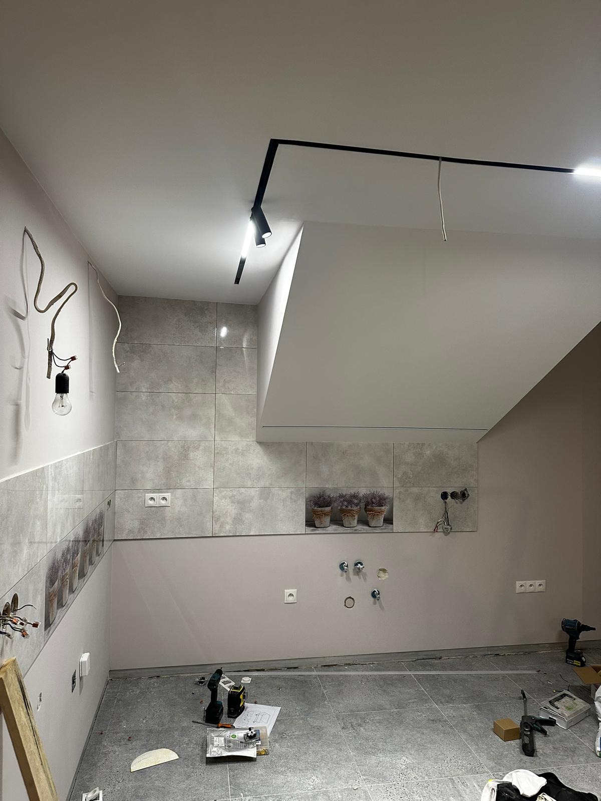 Wnętrze łazienki w trakcie remontu: szare płytki imitujące kamień na ścianach, czarna szyna oświetleniowa z reflektorami na suficie, odsłonięte przewody elektryczne, narzędzia i materiały budowlane...