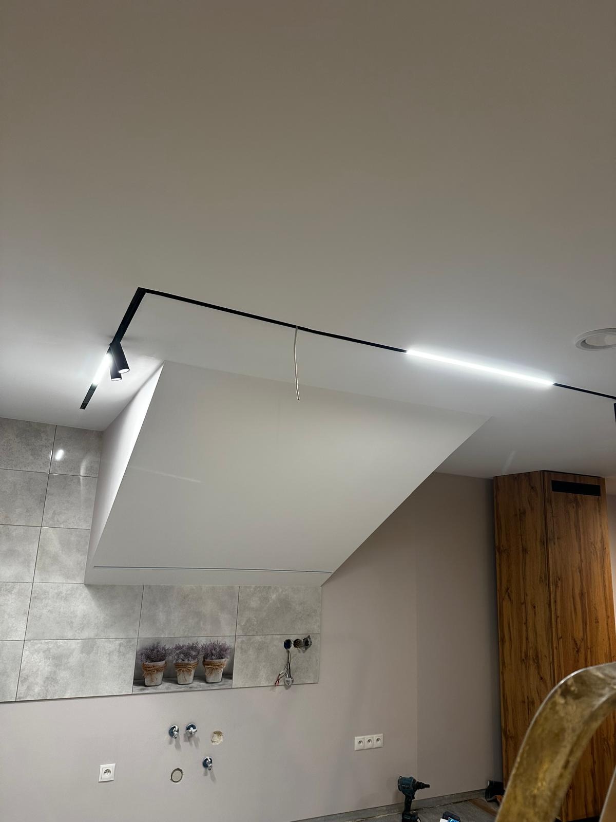 Wnętrze w trakcie prac wykończeniowych: szara płytka na ścianie, czarna szyna oświetleniowa z reflektorami na suficie, wystające kable ze ściany, widoczny fragment drabiny.