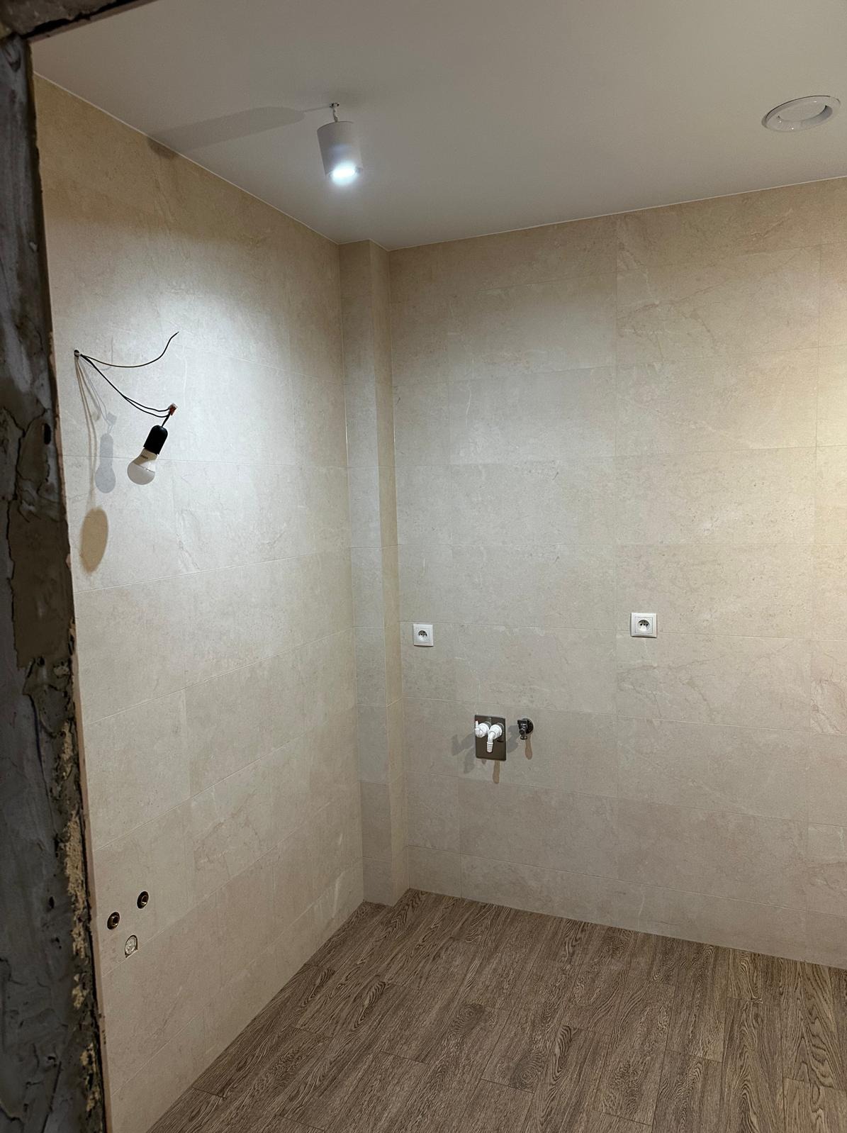 Wnętrze łazienki w trakcie wykańczania: beżowe płytki ścienne, drewnopodobne panele podłogowe, wystające kable z żarówką, gniazdka elektryczne i przyłącze wodne na ścianie.