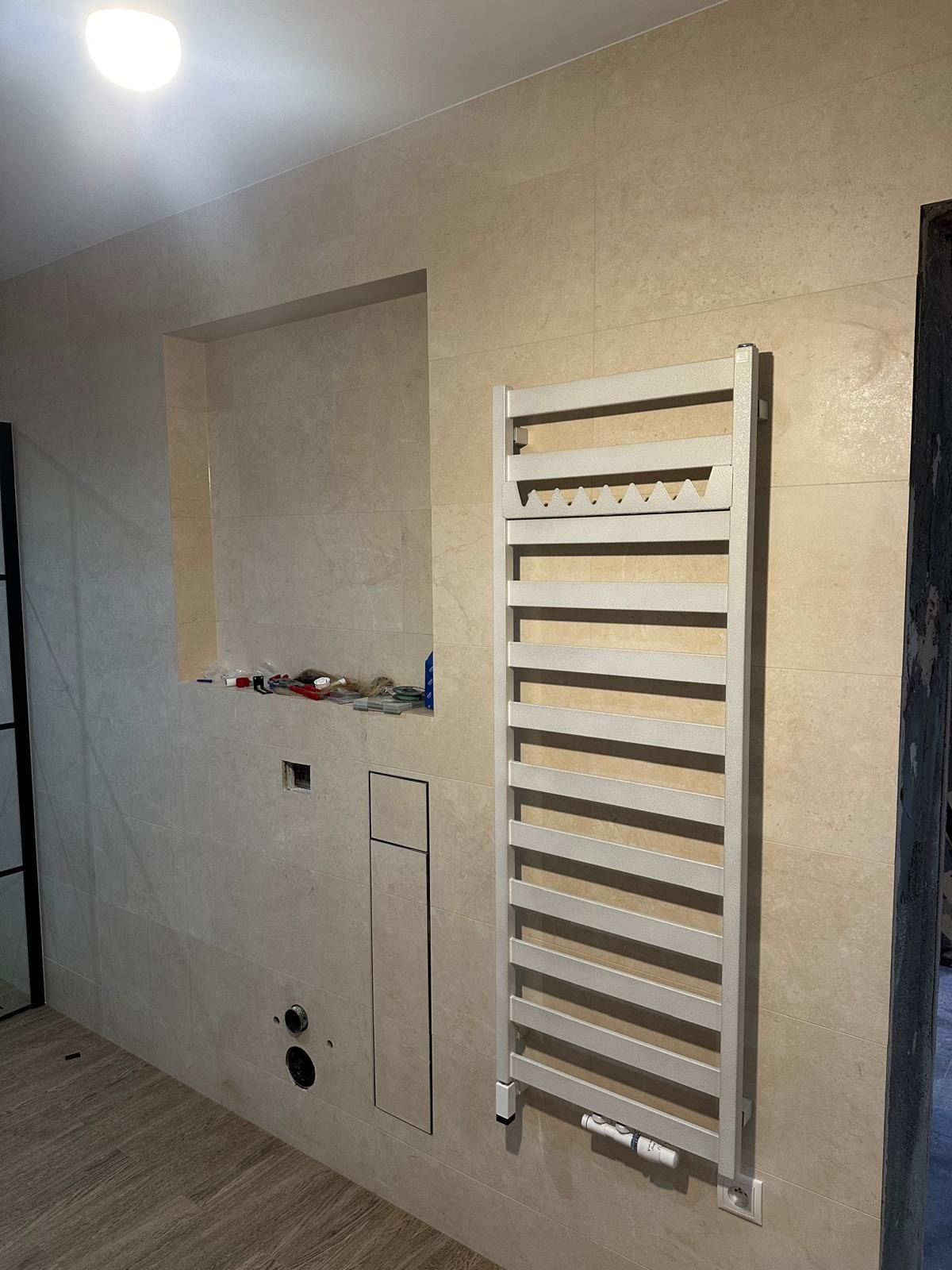 Wnętrze łazienki w trakcie prac wykończeniowych; widoczna ściana z beżowymi płytkami, wnęka z narzędziami, grzejnik drabinkowy i przygotowane podejścia pod armaturę.