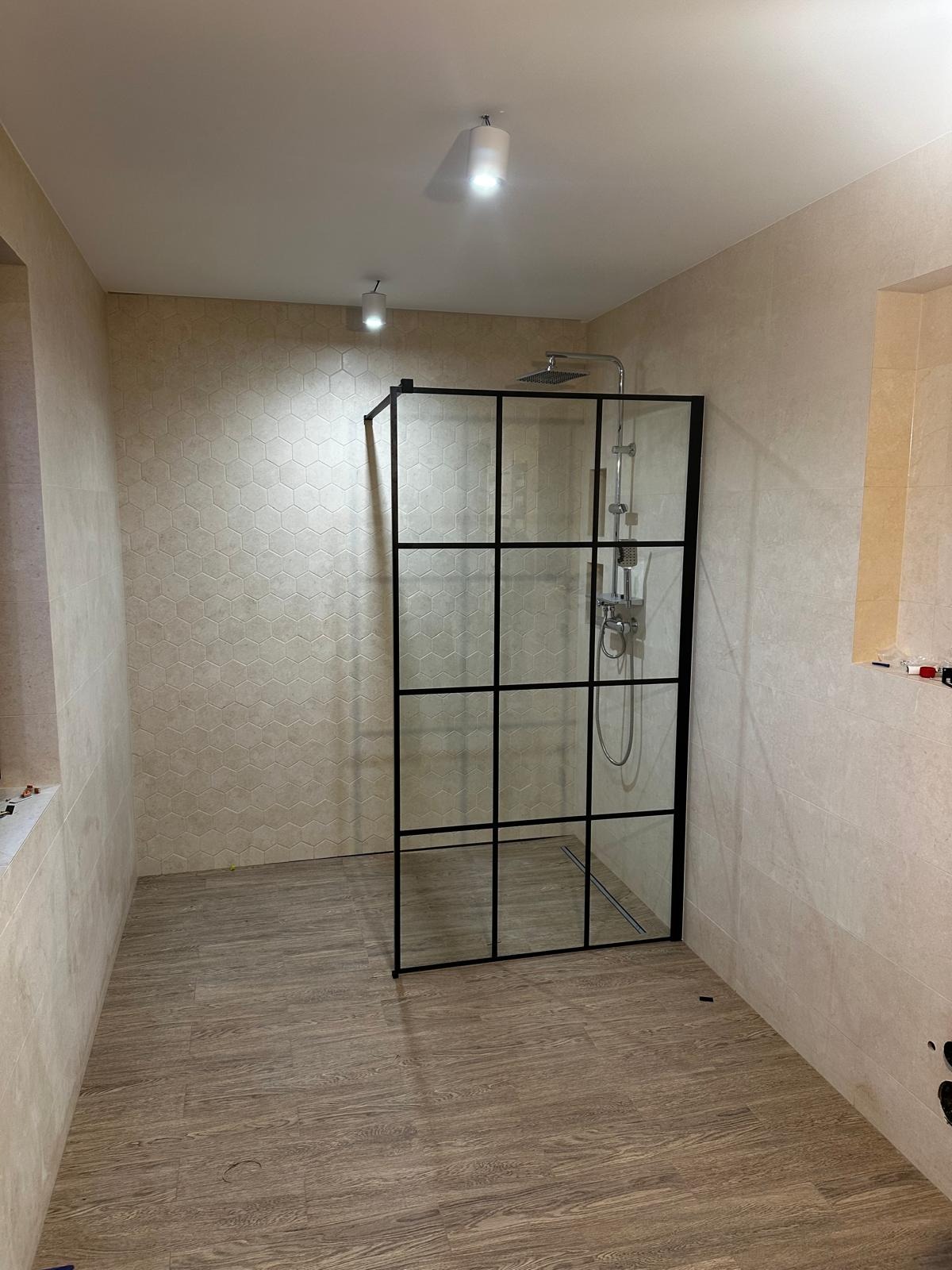 Nowoczesna kabina prysznicowa z czarnymi profilami w łazience z heksagonalnymi płytkami na ścianie i drewnopodobną podłogą, widoczne elementy instalacji elektrycznej.