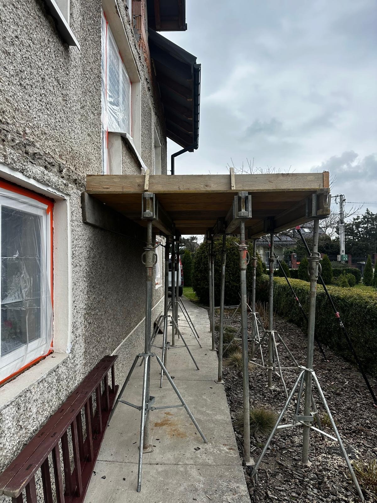 Elewacja domu w trakcie prac przygotowawczych do ocieplenia, widoczne rusztowanie z drewnianym pomostem, okna zabezpieczone folią, drabina oparta o ścianę.