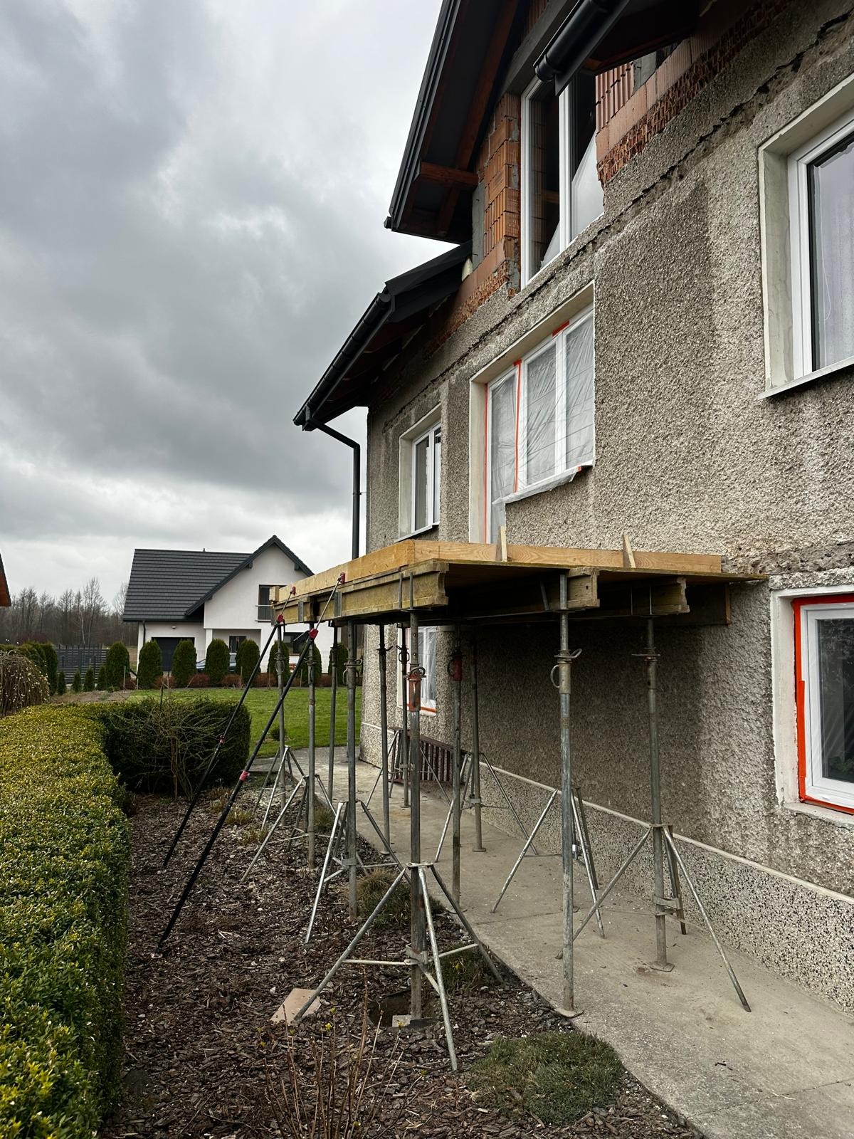 Elewacja domu w trakcie prac remontowych, z widocznym rusztowaniem podporowym i oknami zabezpieczonymi folią; w tle drugi dom i pochmurne niebo.