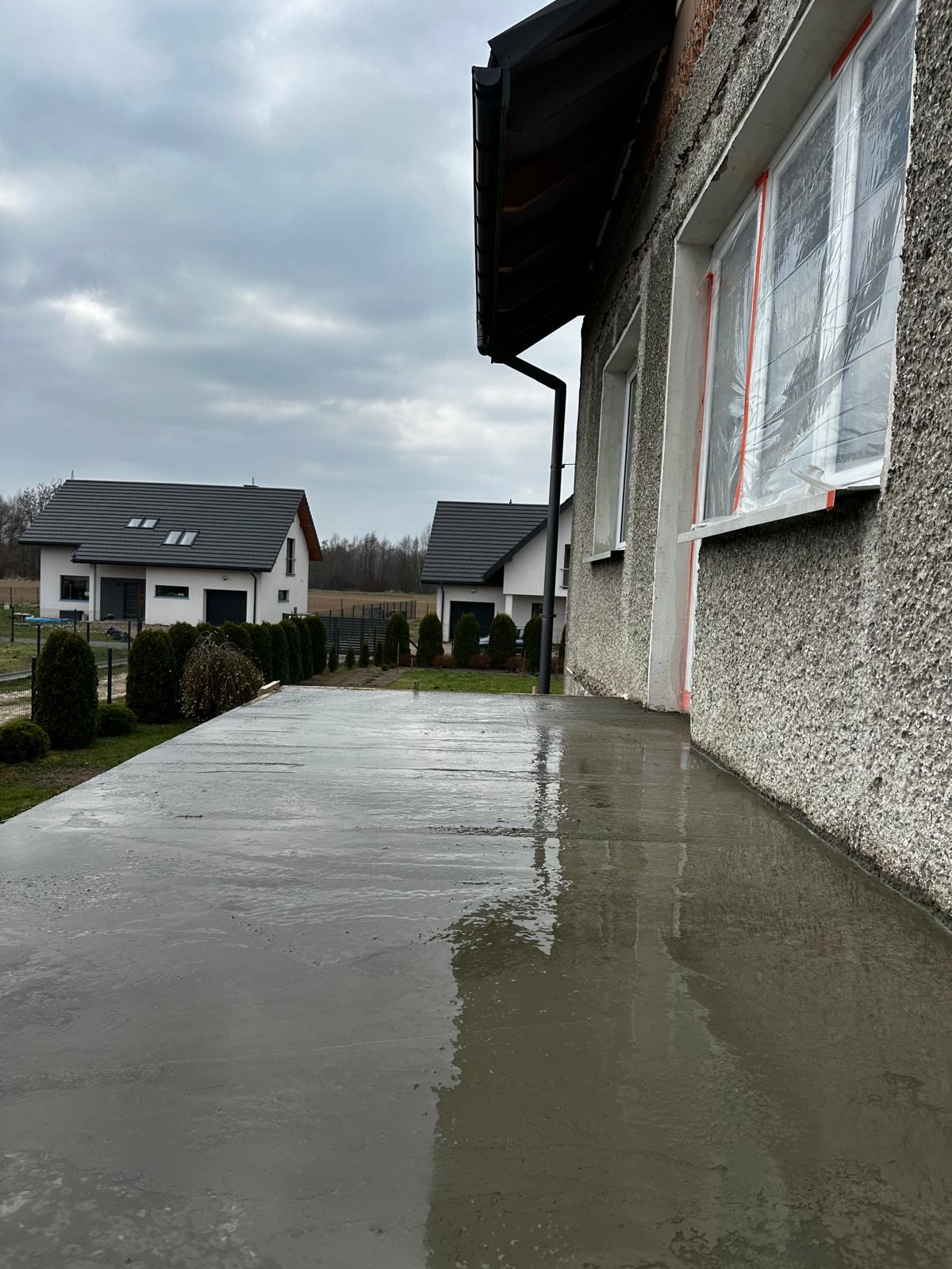 Świeżo wylana, mokra wylewka betonowa tarasu przy szarym domu z oknami zabezpieczonymi folią malarską, w tle widoczne dwa identyczne domy z ciemnymi dachami.