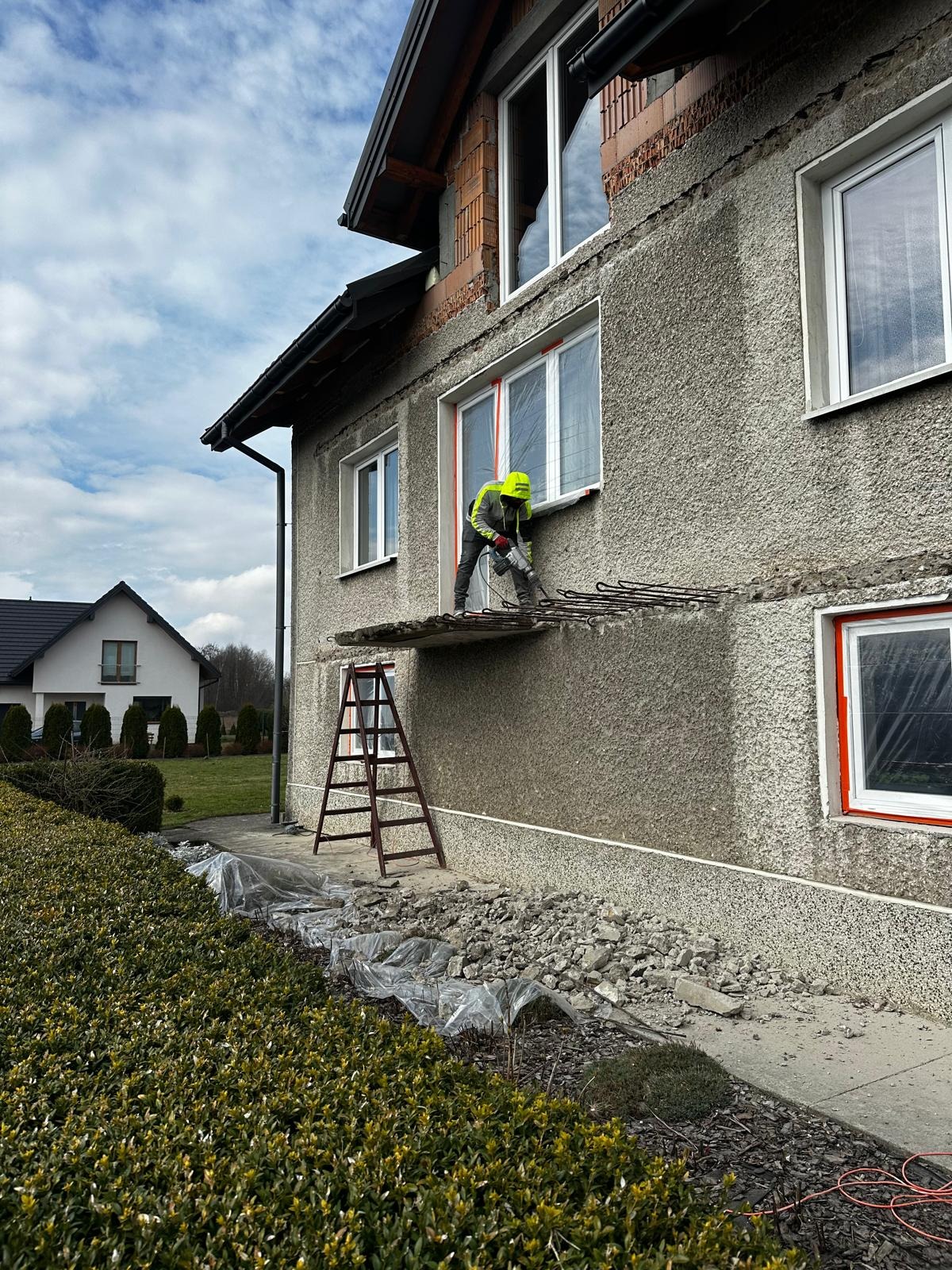 Pracownik w kasku i kamizelce ochronnej skuwa stary tynk z elewacji budynku za pomocą młota pneumatycznego, widoczne rusztowanie, gruz i folia zabezpieczająca.