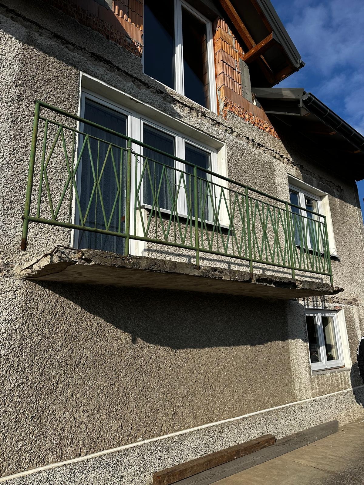 Niezabezpieczony balkon z zieloną, metalową balustradą, z widocznymi uszkodzeniami betonu, na tle szarej, chropowatej elewacji budynku z odkrytą cegłą i oknami.