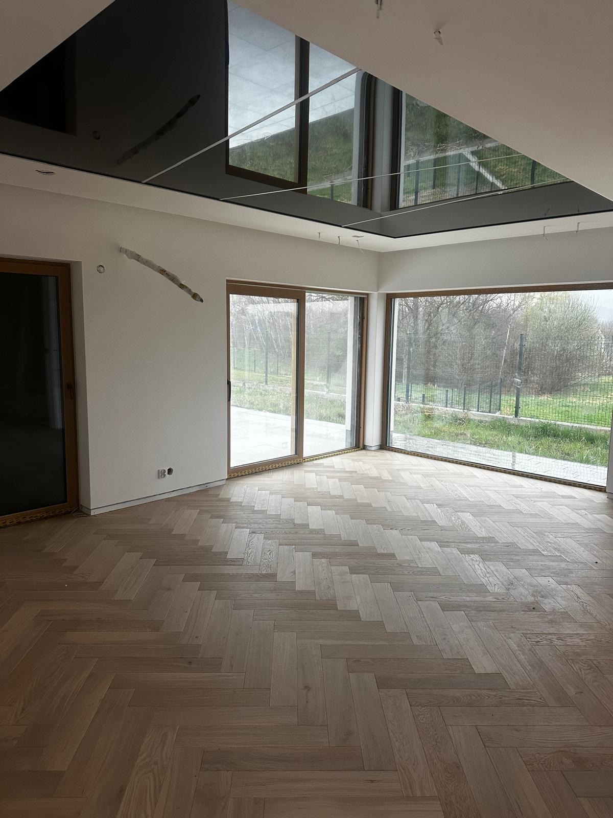 Jasny parkiet ułożony w jodełkę francuską w przestronnym, minimalistycznym wnętrzu z dużymi oknami i czarnym, lustrzanym sufitem.