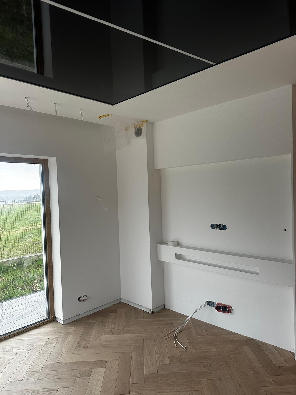 Wnętrze pokoju w trakcie prac wykończeniowych: parkiet w jodełkę, białe ściany z przygotowanymi gniazdkami elektrycznymi, czarny, lustrzany sufit podwieszany, zabezpieczone elementy instalacji.