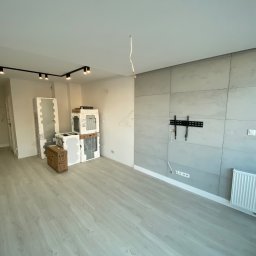 PerfectHome - Wnętrze pokoju w trakcie wykańczania: szare panele ścienne, zamontowany uchwyt TV, grzejnik, podłoga z paneli, oświetlenie sufitowe na szynie i kable zwisające z sufitu.