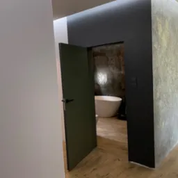 Otwarta, ciemnozielona łazienkowa prowadząca do łazienki z wolnostojącą wanną na tle ściany z efektem rdzy, widok z korytarza z podłogą imitującą drewno.