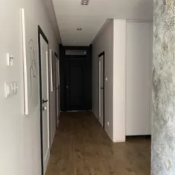 Długi korytarz z drewnopodobną podłogą, białymi ścianami z czarnymi ramami drzwi i abstrakcyjnymi obrazami, dekoracyjną ścianą z efektem betonu i wzorem w rogu, oraz wbudowaną białą szafą.