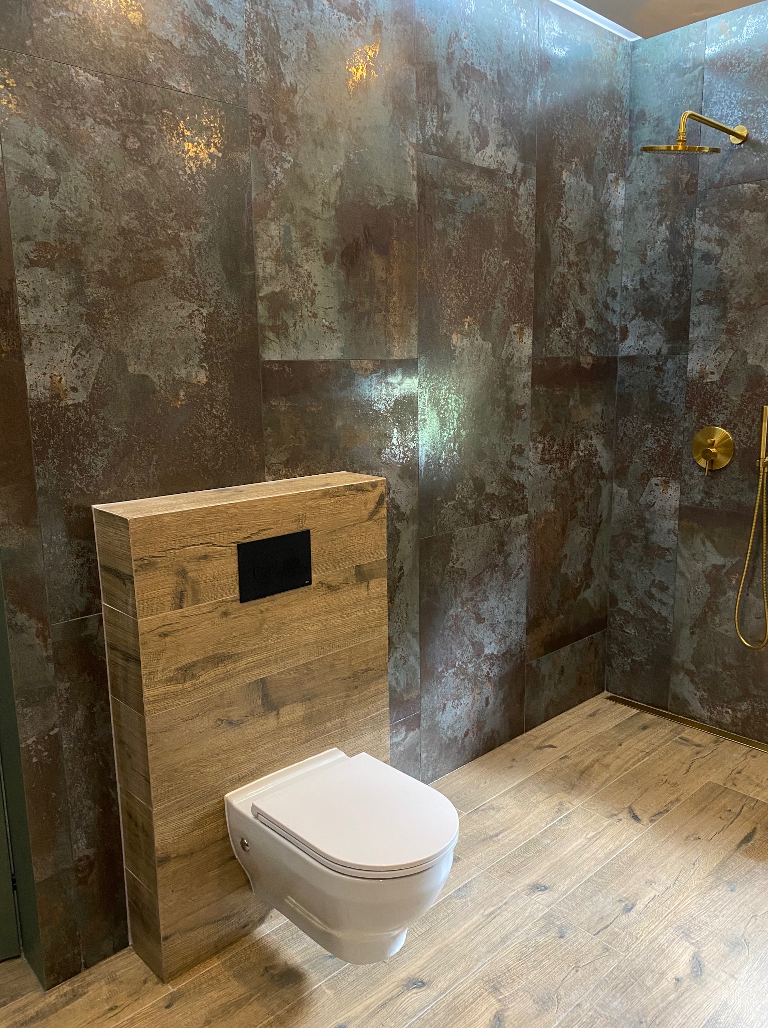 Nowoczesna łazienka z wiszącą toaletą zabudowaną drewnopodobną płytą, ciemnymi płytkami imitującymi rdzę na ścianach i złotą armaturą prysznicową.