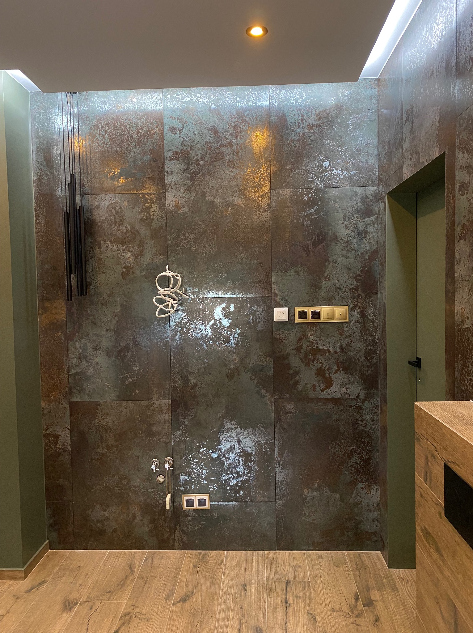 Ściana wykończona płytkami imitującymi rdzę, z widocznymi elementami instalacji elektrycznej i wodnej przed zabudową, oświetlenie sufitowe LED, podłoga wyłożona panelami drewnopodobnymi.