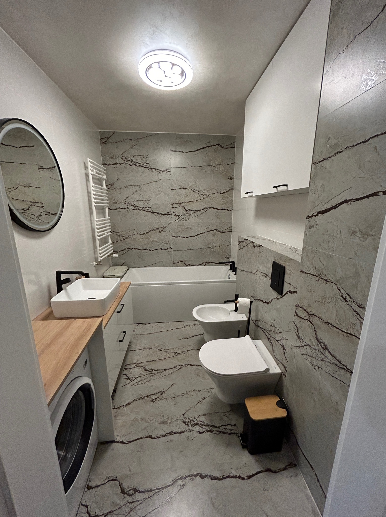 Nowoczesna łazienka w Radomsku z marmurowymi płytkami, umywalką nablatową z drewnianym blatem, wiszącą toaletą i bidetem oraz pralką zabudowaną pod blatem.