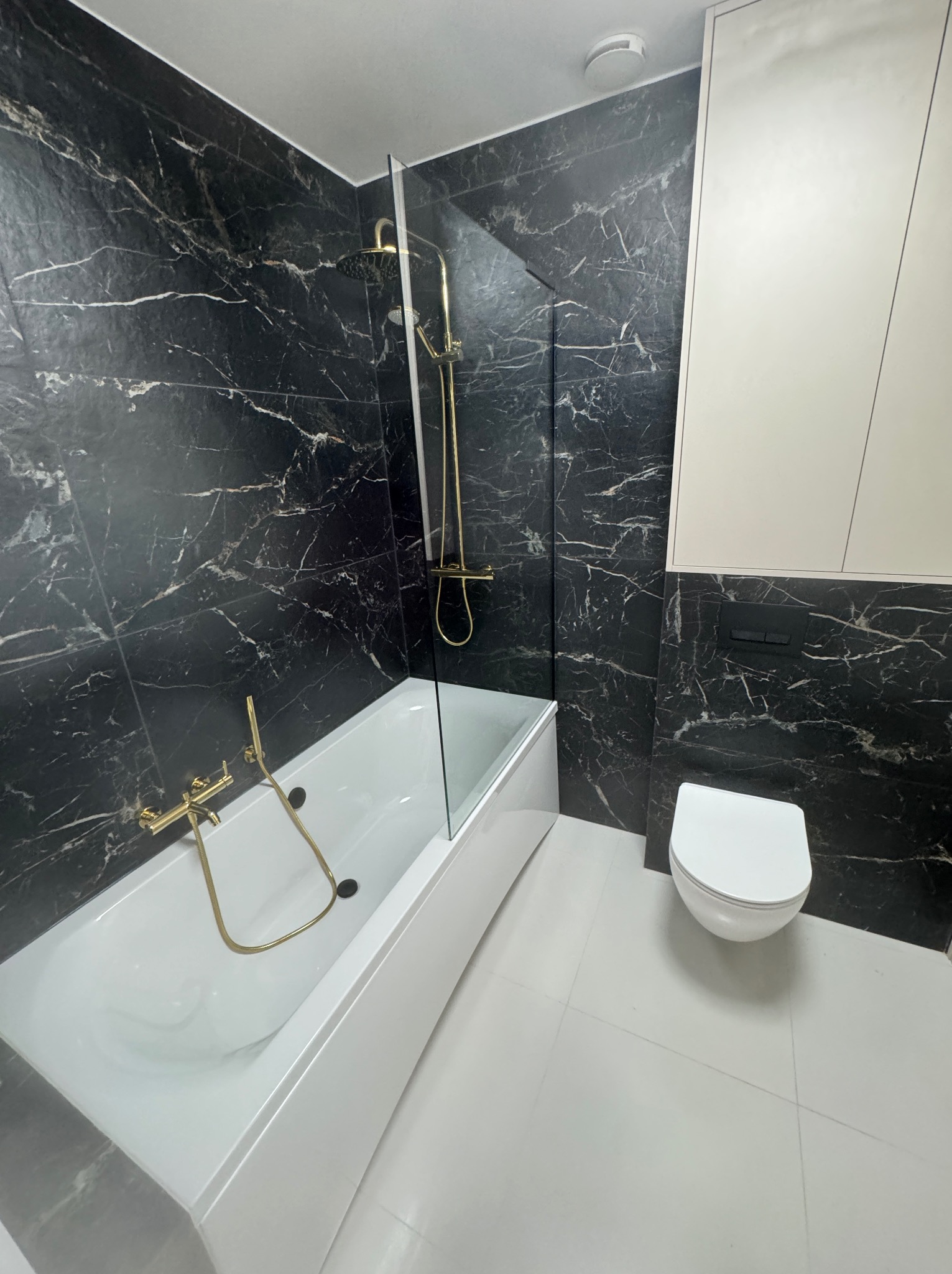 Nowoczesna łazienka z czarnymi płytkami imitującymi marmur, złotą armaturą i białą ceramiką sanitarną. Kabina prysznicowa ze szklaną ścianką. Widoczne białe szafki wiszące.