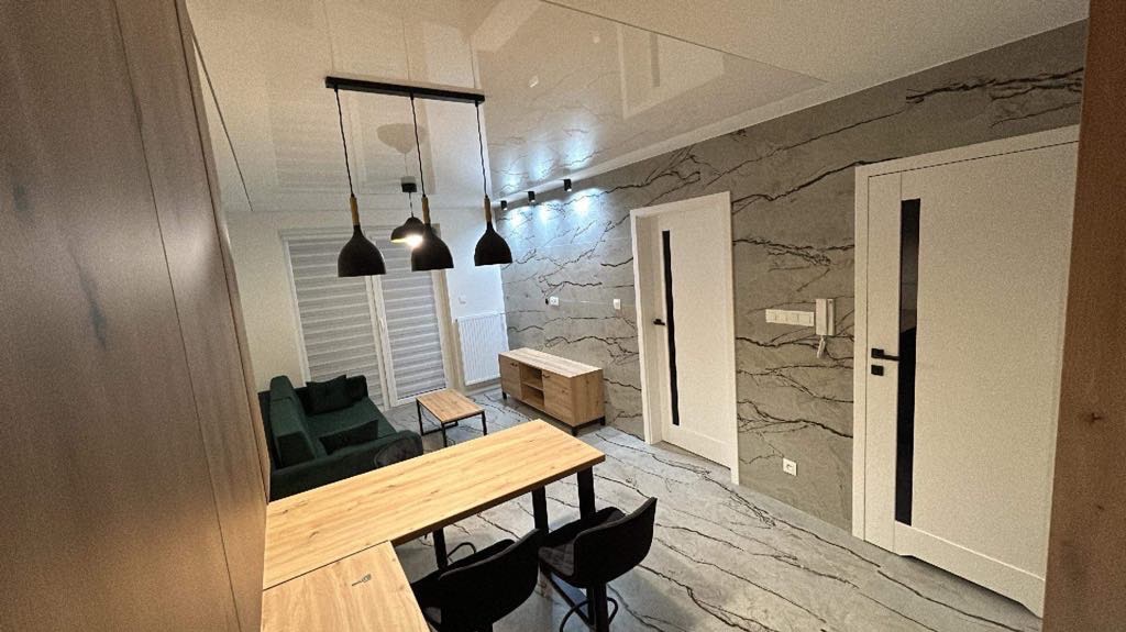 Nowoczesne wnętrze salonu z błyszczącym sufitem, szarą ścianą imitującą marmur, drewnianym stołem z krzesłami barowymi i designerskimi lampami wiszącymi.