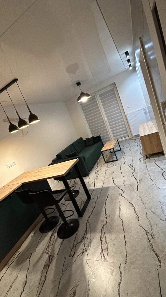 Nowoczesne wnętrze mieszkania z błyszczącym, białym sufitem i podłogą imitującą marmur z wyraźnym wzorem żyłkowania, zielona sofa narożna i drewniany blat baru z czarnymi hokerami.