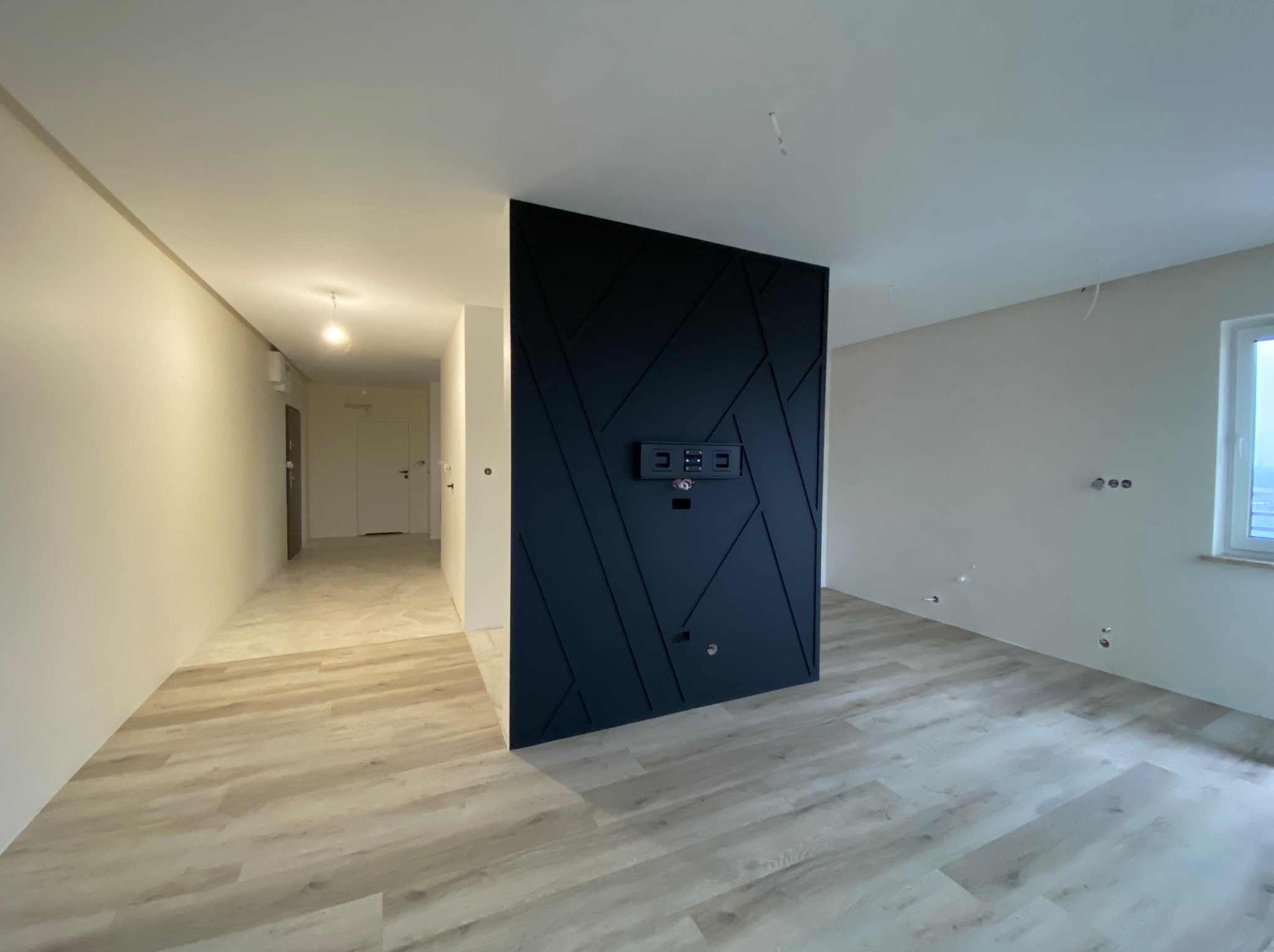 Nowoczesne wnętrze: czarna ściana działowa z geometrycznym wzorem i uchwytem na telewizor, panele podłogowe w odcieniach beżu, białe ściany i sufit, okno z widokiem na zewnątrz.