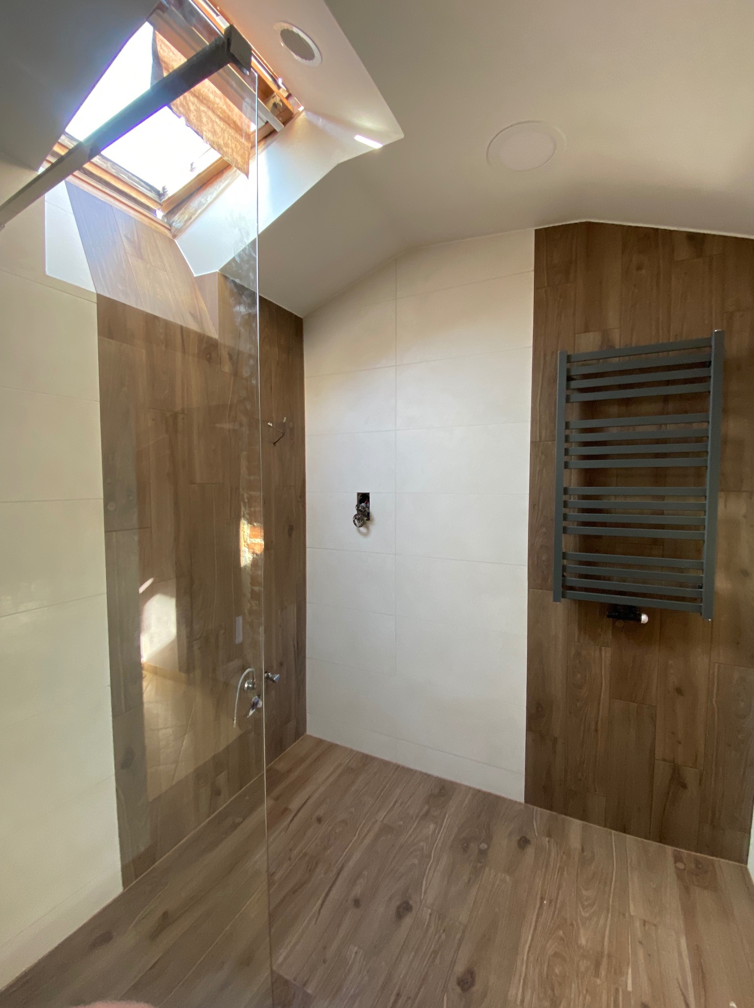 Nowoczesna łazienka na poddaszu z drewnopodobnymi płytkami na podłodze i ścianach, kabiną prysznicową ze szklaną ścianką i grzejnikiem drabinkowym.