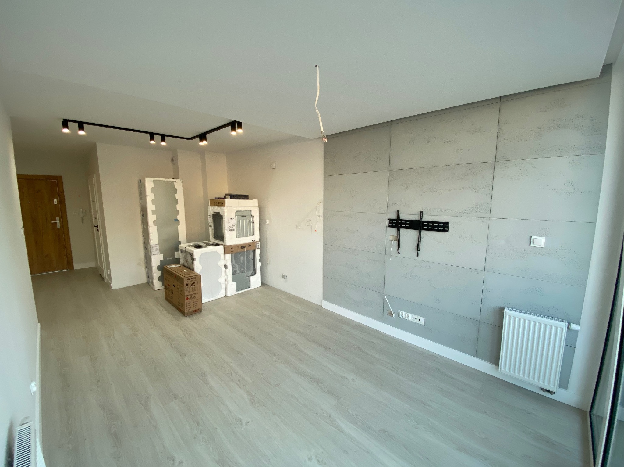 Wnętrze pokoju w trakcie wykańczania: szare panele ścienne, zamontowany uchwyt TV, grzejnik, podłoga z paneli, oświetlenie sufitowe na szynie i kable zwisające z sufitu.