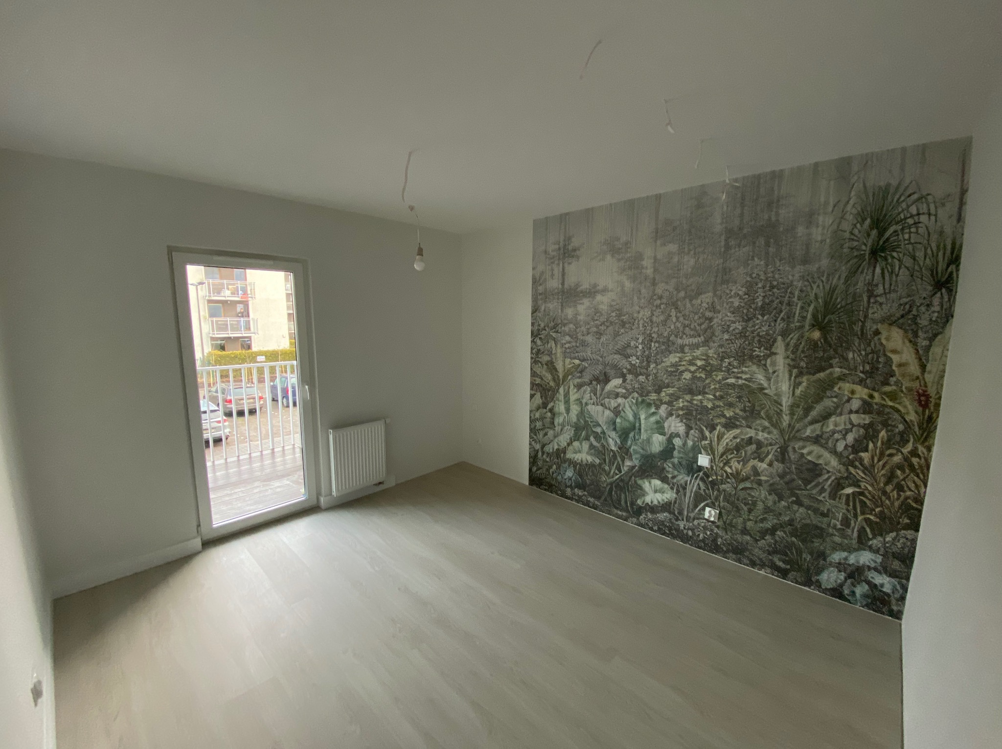 Pusty pokój z jasnymi panelami podłogowymi i fototapetą z motywem dżungli na ścianie, widok na balkon przez otwarte drzwi balkonowe, surowe wykończenie sufitu z wystającymi kablami.