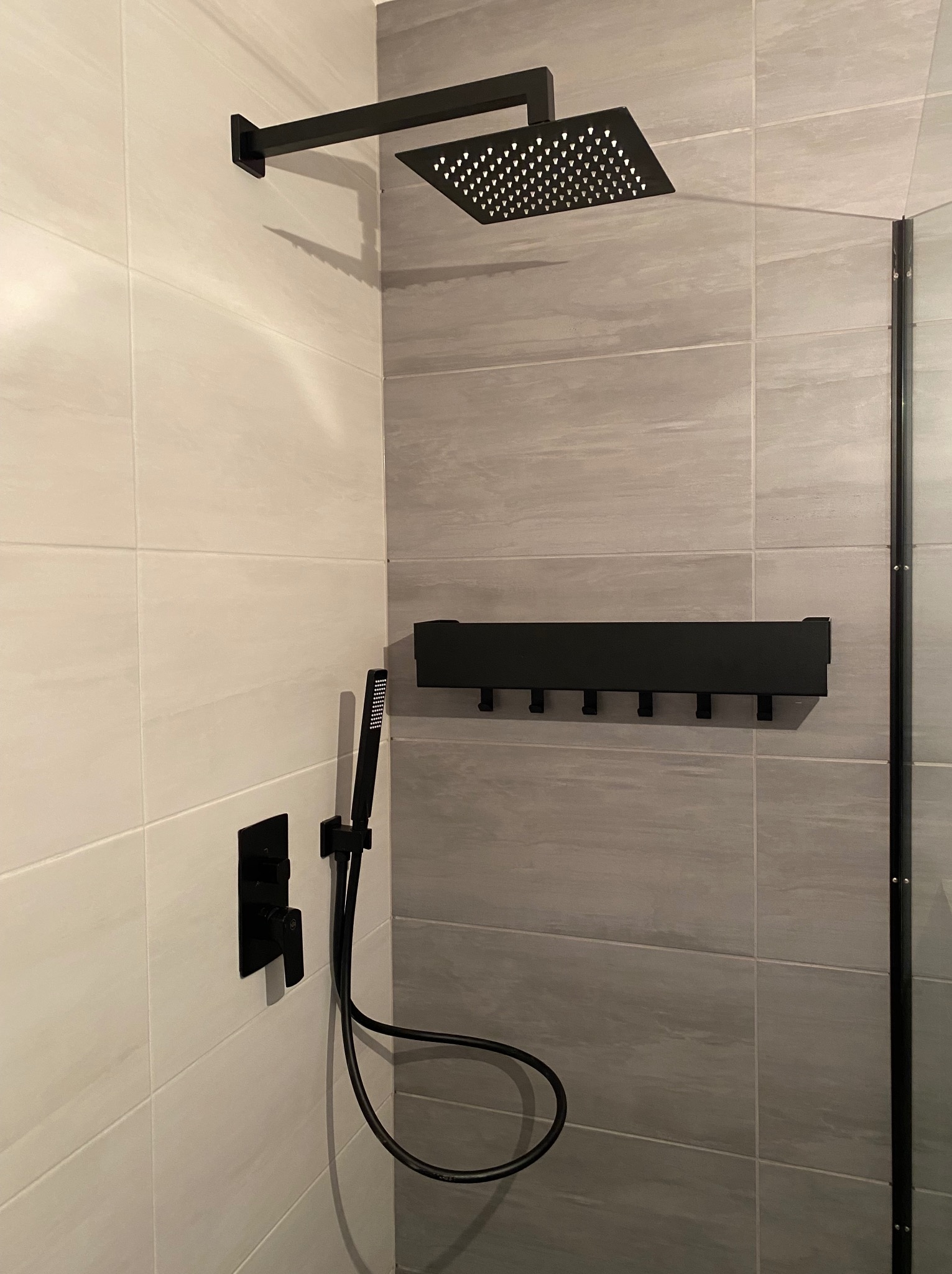Nowoczesna kabina prysznicowa z czarną armaturą: deszczownica, słuchawka prysznicowa, bateria podtynkowa i wieszak na ręczniki na tle płytek imitujących kamień.