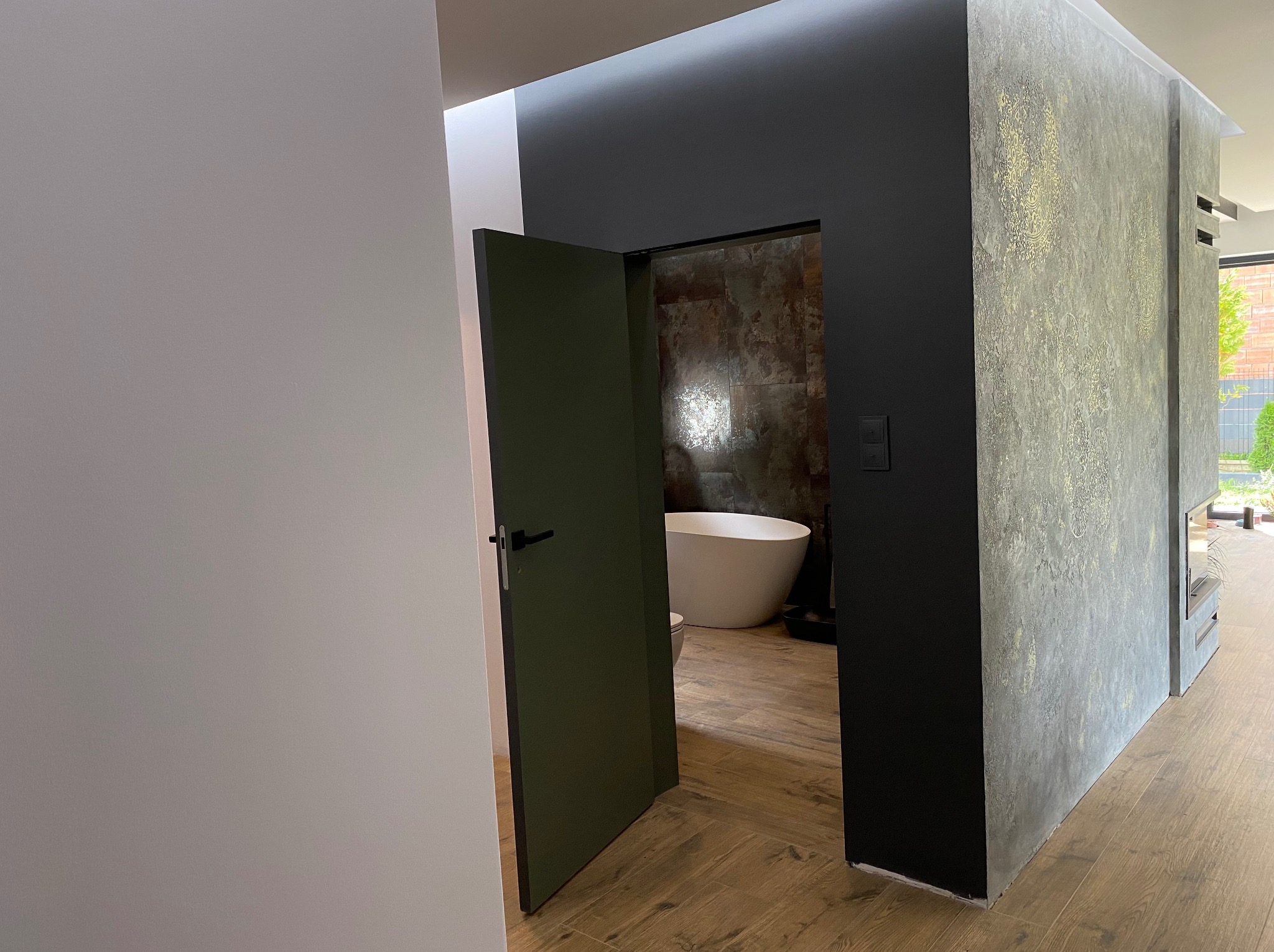 Otwarta, ciemnozielona łazienkowa prowadząca do łazienki z wolnostojącą wanną na tle ściany z efektem rdzy, widok z korytarza z podłogą imitującą drewno.