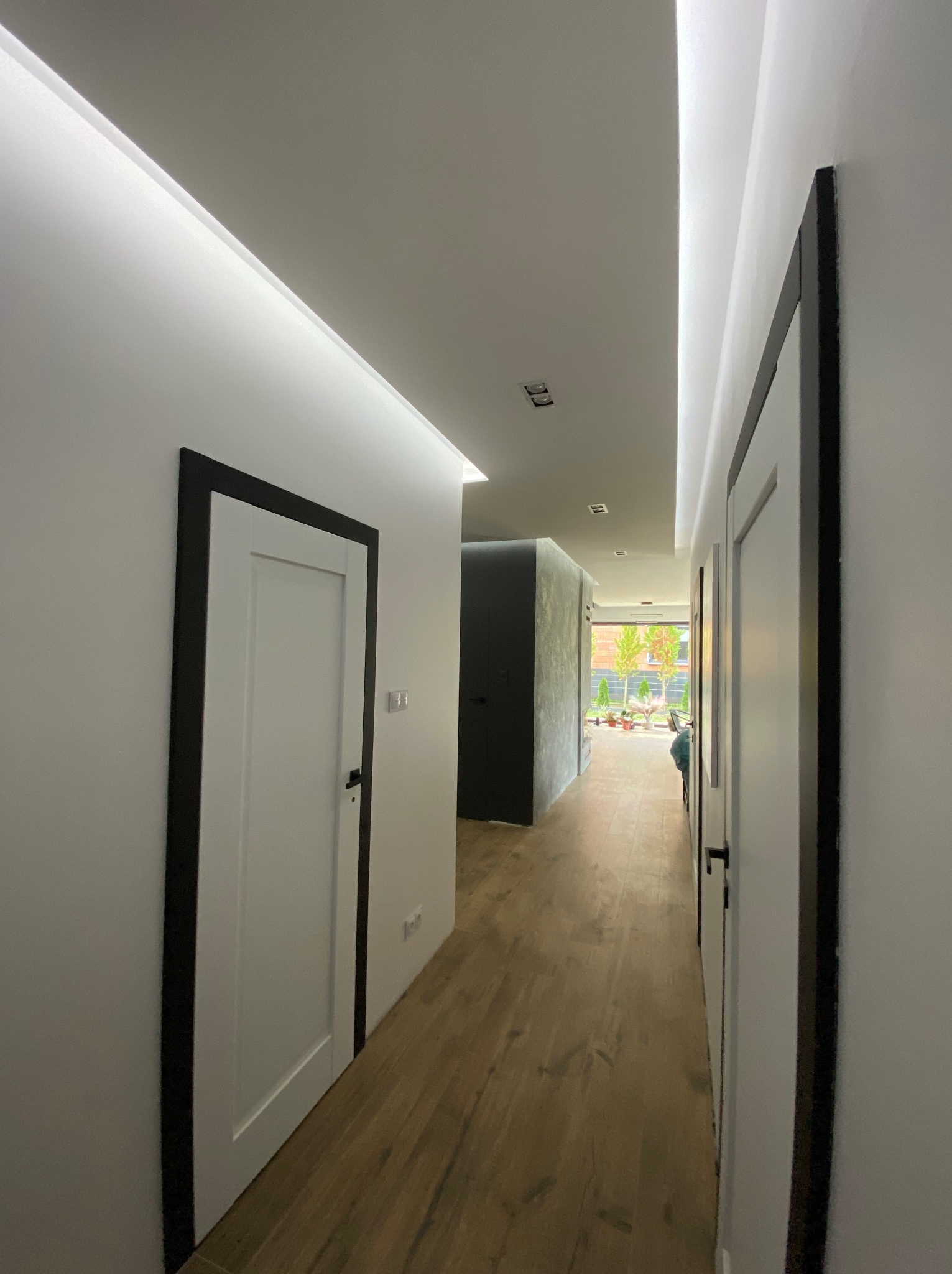 Nowoczesny korytarz z białymi ścianami, drewnianą podłogą i białymi drzwiami z czarnymi ościeżnicami, oświetlenie LED w suficie podwieszanym.