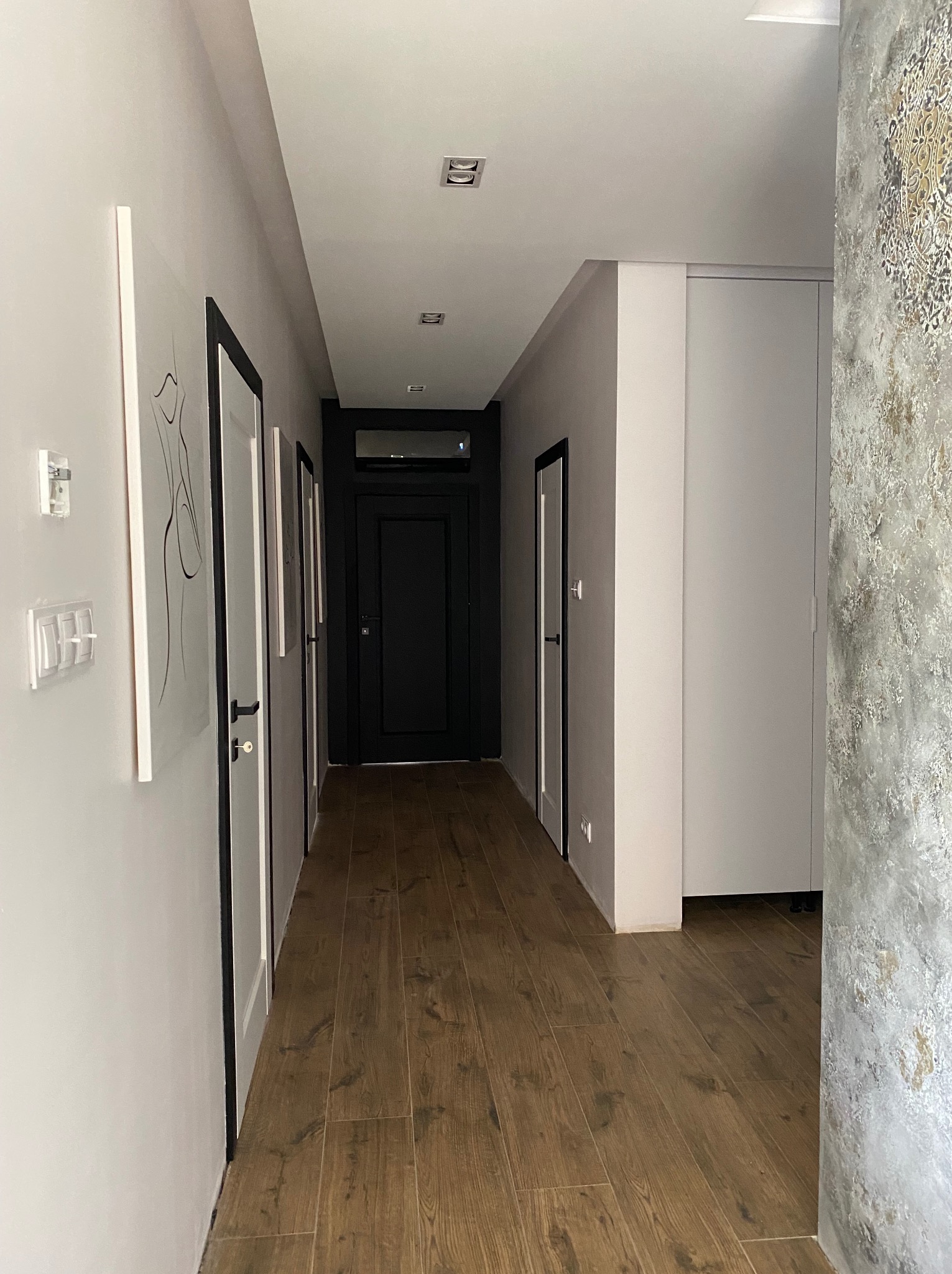 Długi korytarz z drewnopodobną podłogą, białymi ścianami z czarnymi ramami drzwi i abstrakcyjnymi obrazami, dekoracyjną ścianą z efektem betonu i wzorem w rogu, oraz wbudowaną białą szafą.