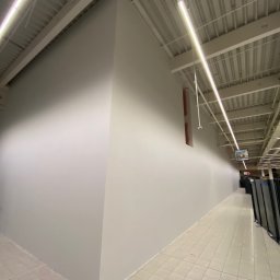 LEO_BUD VOLODYMYR KOZLOVSKYY - Wnętrze komercyjne po malowaniu: gładkie, szare ściany wzdłuż korytarza, oświetlone lampami sufitowymi. Widoczna podłoga z płytek i fragment regałów w oddali.