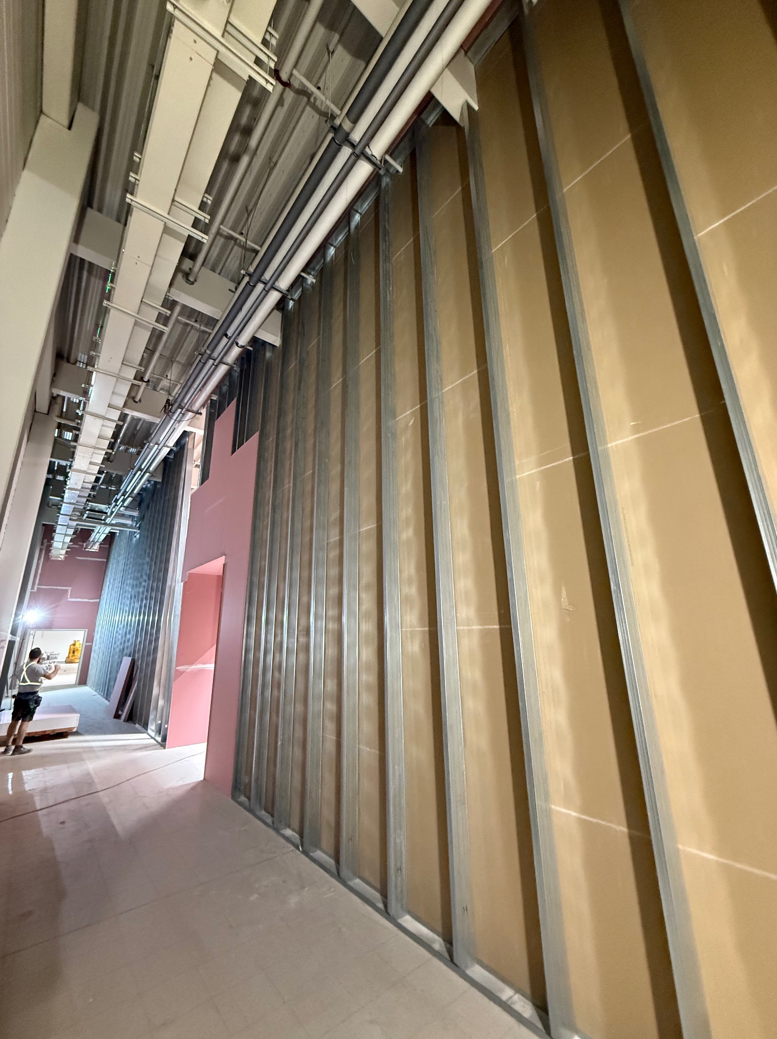 Wnętrze w trakcie prac: metalowy stelaż pod zabudowę z płyt, widoczne instalacje pod sufitem, fragment różowej izolacji, pracownik w oddali. Surowy stan wykończenia.