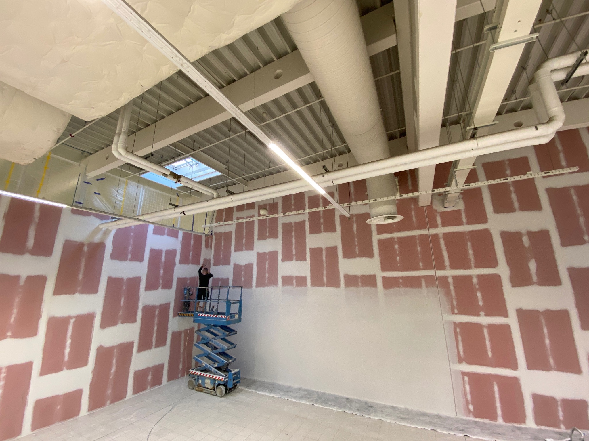 Wnętrze w trakcie prac wykończeniowych: częściowo wyszpachlowane ściany na różowo, pracownik na podnośniku. Widoczne instalacje pod sufitem.