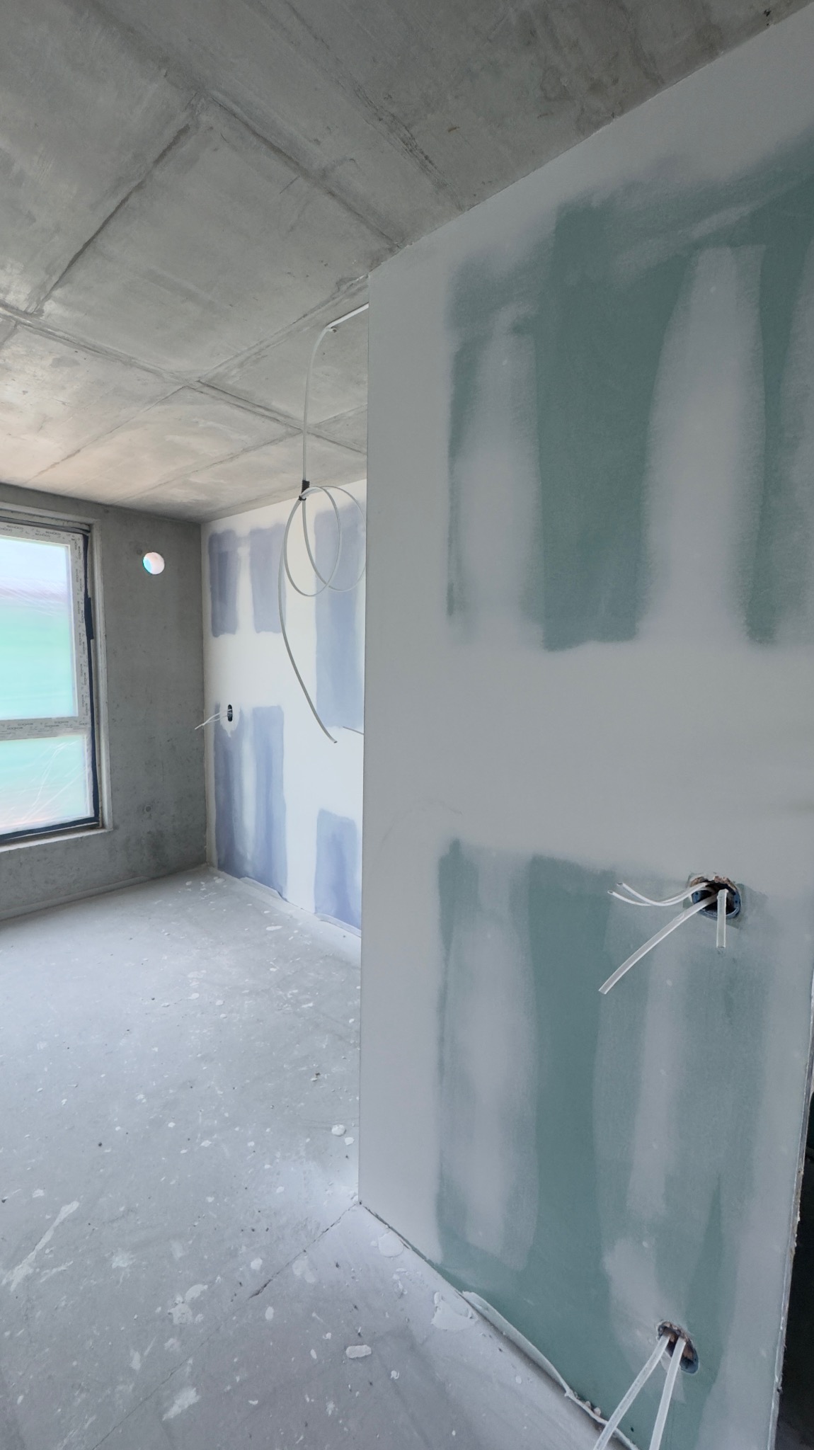 Surowe wnętrze w trakcie wykańczania: ściany z płyt gipsowo-kartonowych, częściowo szpachlowane, widoczne kable elektryczne, betonowy sufit i okno. Etap prac wykończeniowych.