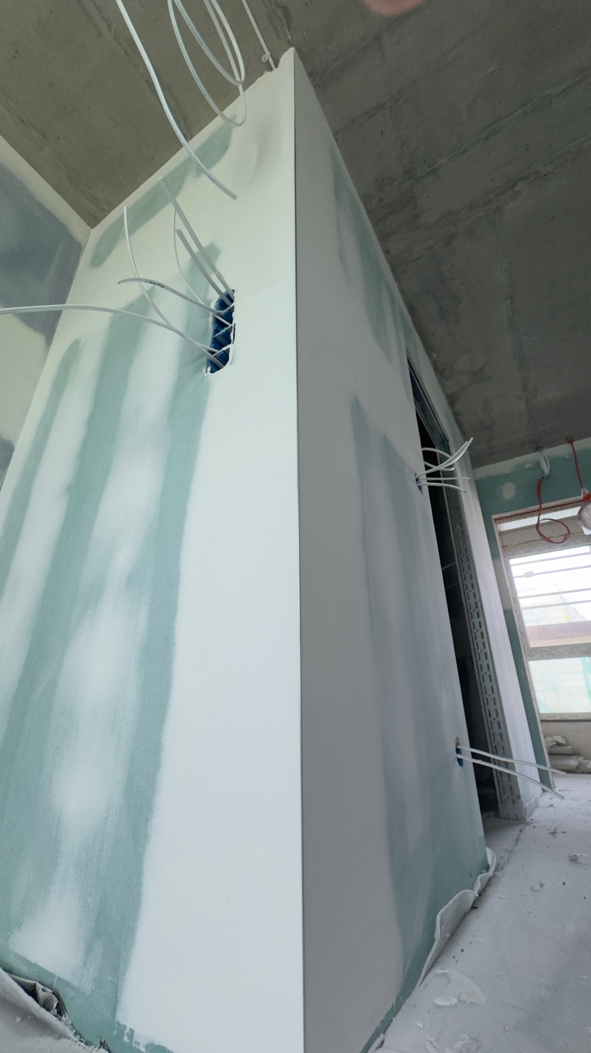 Wnętrze w trakcie wykańczania: ściany z płyt gipsowych, częściowo pokryte szpachlą, widoczne przewody elektryczne i surowy betonowy sufit. Ujęcie z dołu.