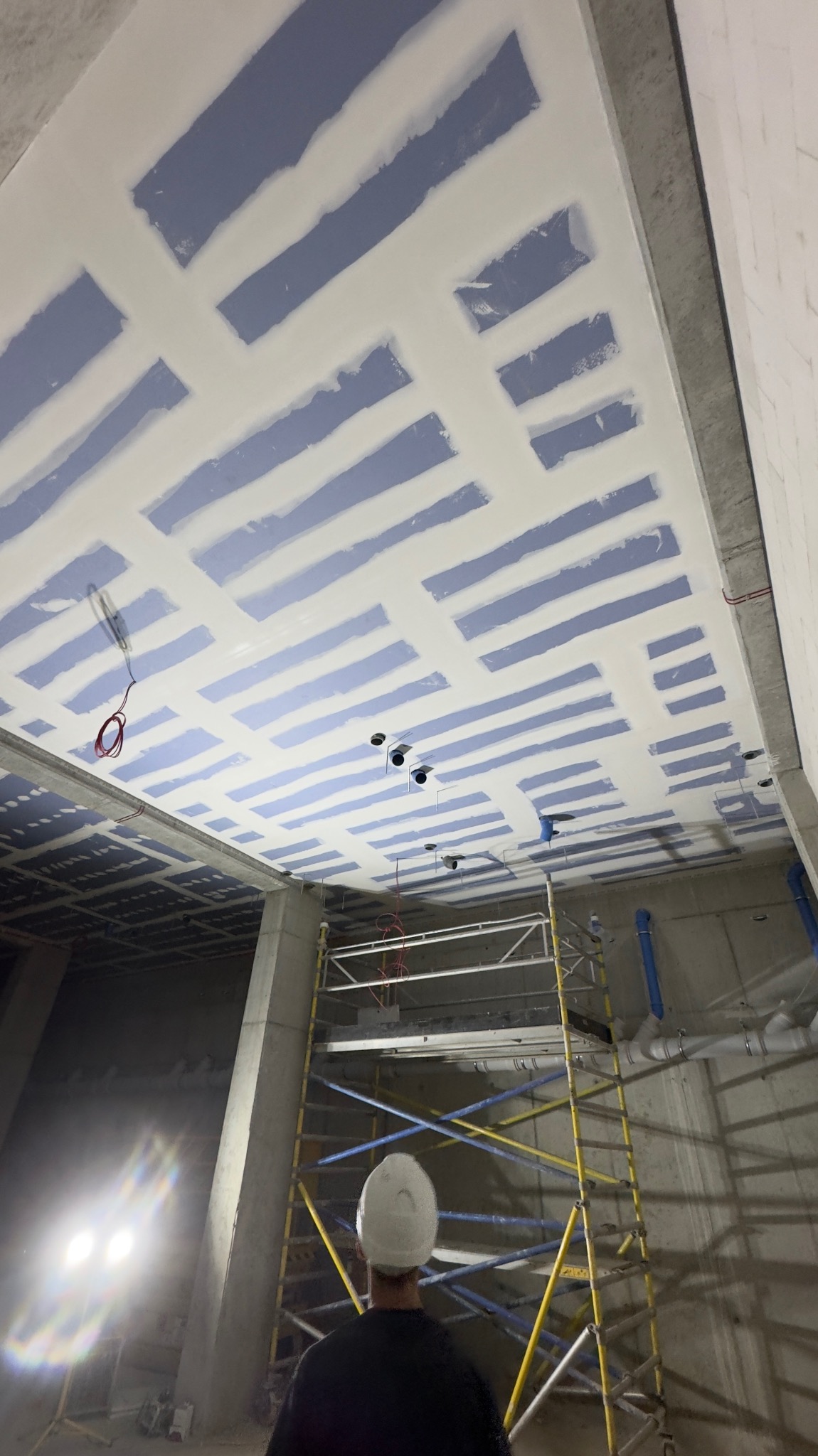 Sufit z płyt karton-gips, częściowo wyszpachlowany, z widocznymi instalacjami i rusztowaniem. Pracownik w kasku obserwuje postęp prac wykończeniowych.