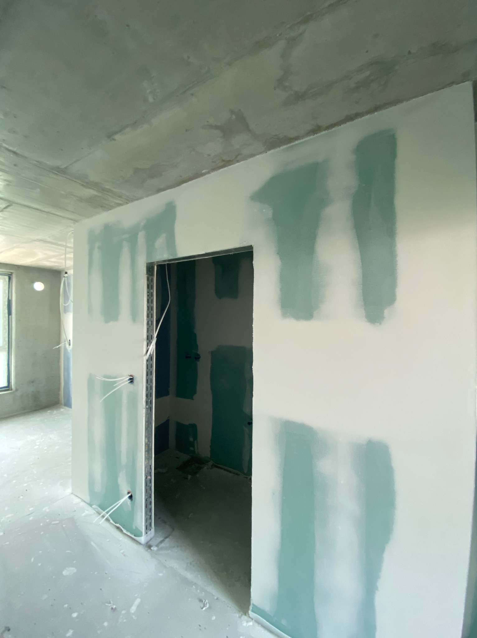 Wnętrze w trakcie prac wykończeniowych: ściany z płyt gipsowych z widocznymi miejscami szpachlowania, surowy betonowy sufit, widok na okno i instalacje elektryczne.