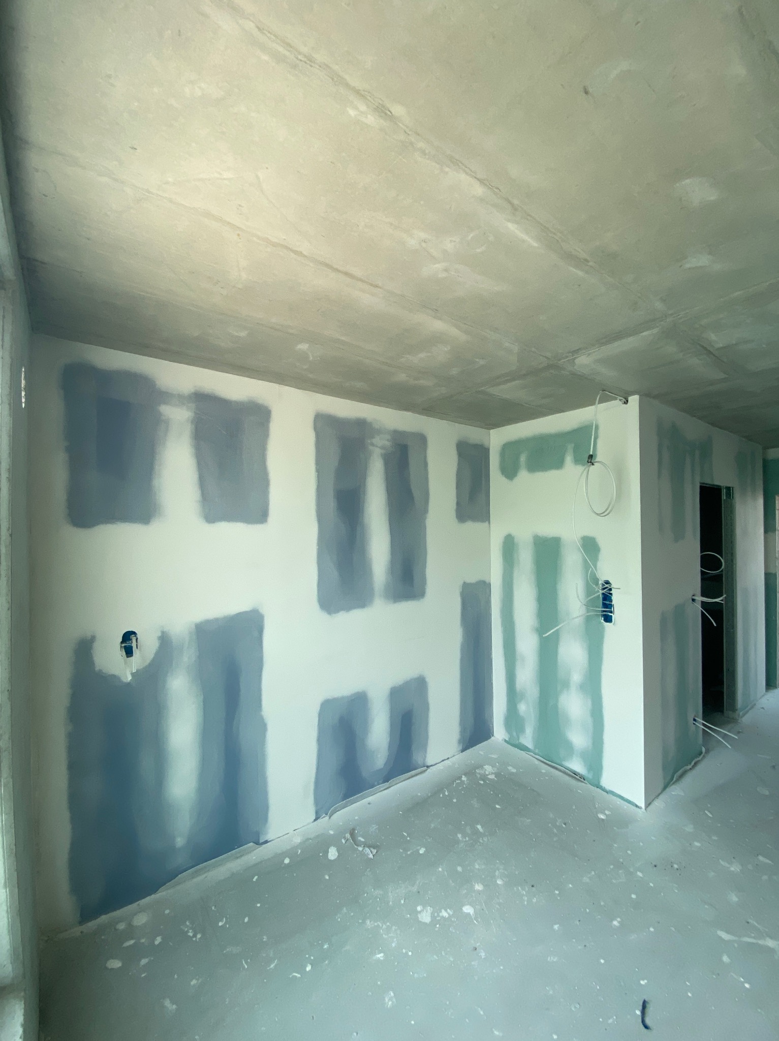 Wnętrze w trakcie prac: ściany po wstępnym szpachlowaniu, widoczne fragmenty instalacji elektrycznej. Surowy betonowy sufit, przygotowanie do dalszych etapów wykończenia.