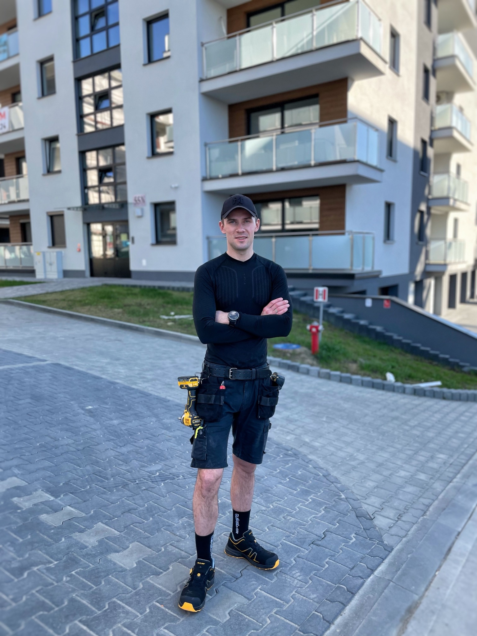Młody mężczyzna w stroju roboczym z narzędziami na tle nowoczesnego budynku mieszkalnego. Uśmiechnięty, z założonymi rękami. Widoczne detale wyposażenia.
