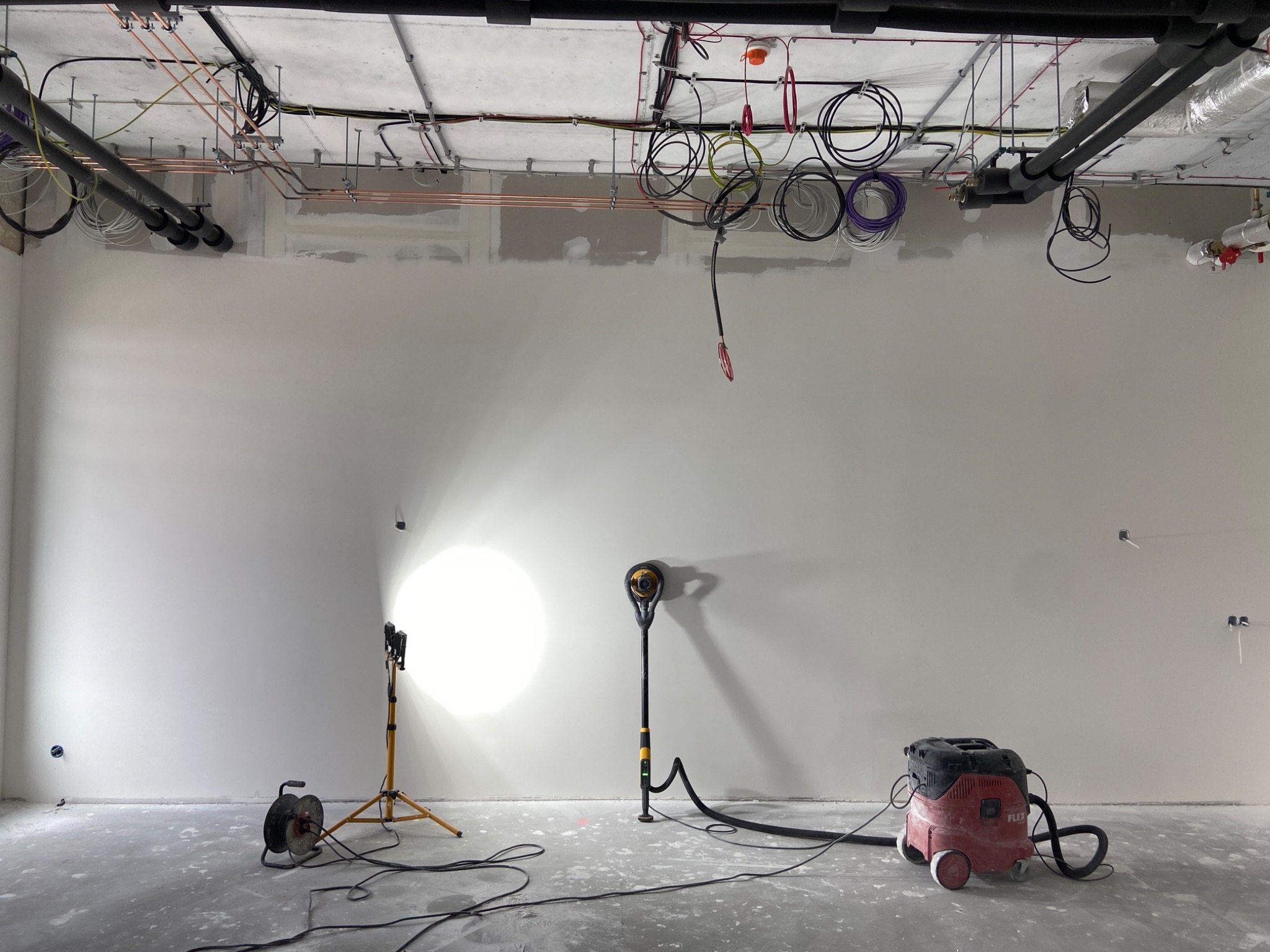 Wnętrze w trakcie prac wykończeniowych: gładka, jasna ściana oświetlona lampą budowlaną, szlifierka do gipsu i odkurzacz przemysłowy. Widoczne instalacje pod sufitem.