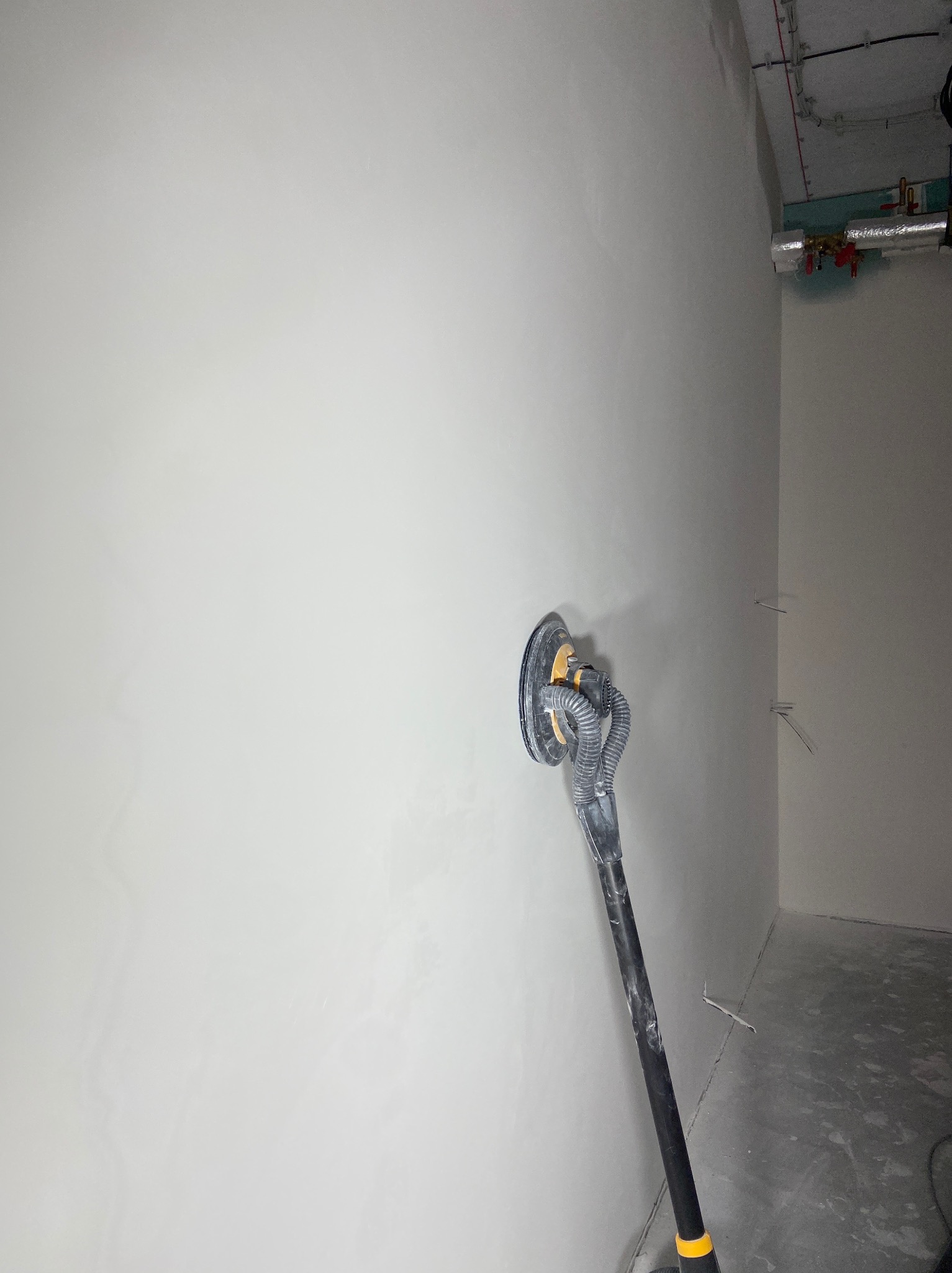 Szlifowanie ściany szlifierką z odkurzaczem w rogu pomieszczenia. Widoczne przewody i rury pod sufitem. Ściana przygotowana do dalszych prac wykończeniowych.