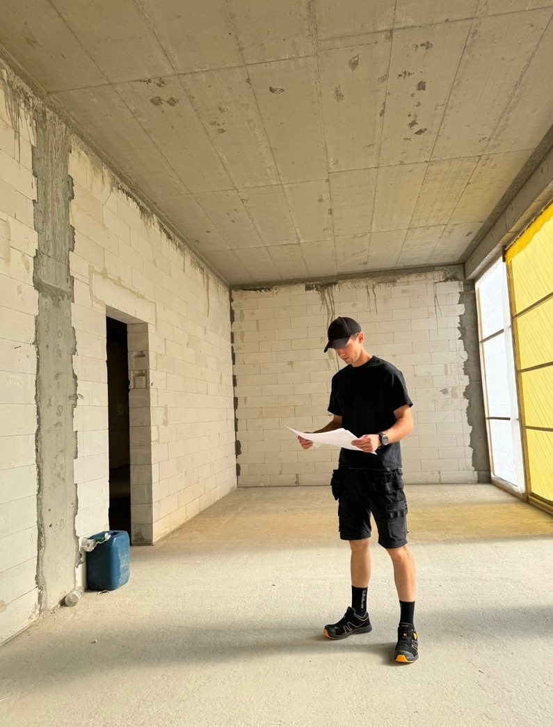 Mężczyzna w czarnym stroju roboczym analizuje plan w surowym wnętrzu z białych bloczków, z widocznymi śladami prac budowlanych i oknami z żółtą folią.