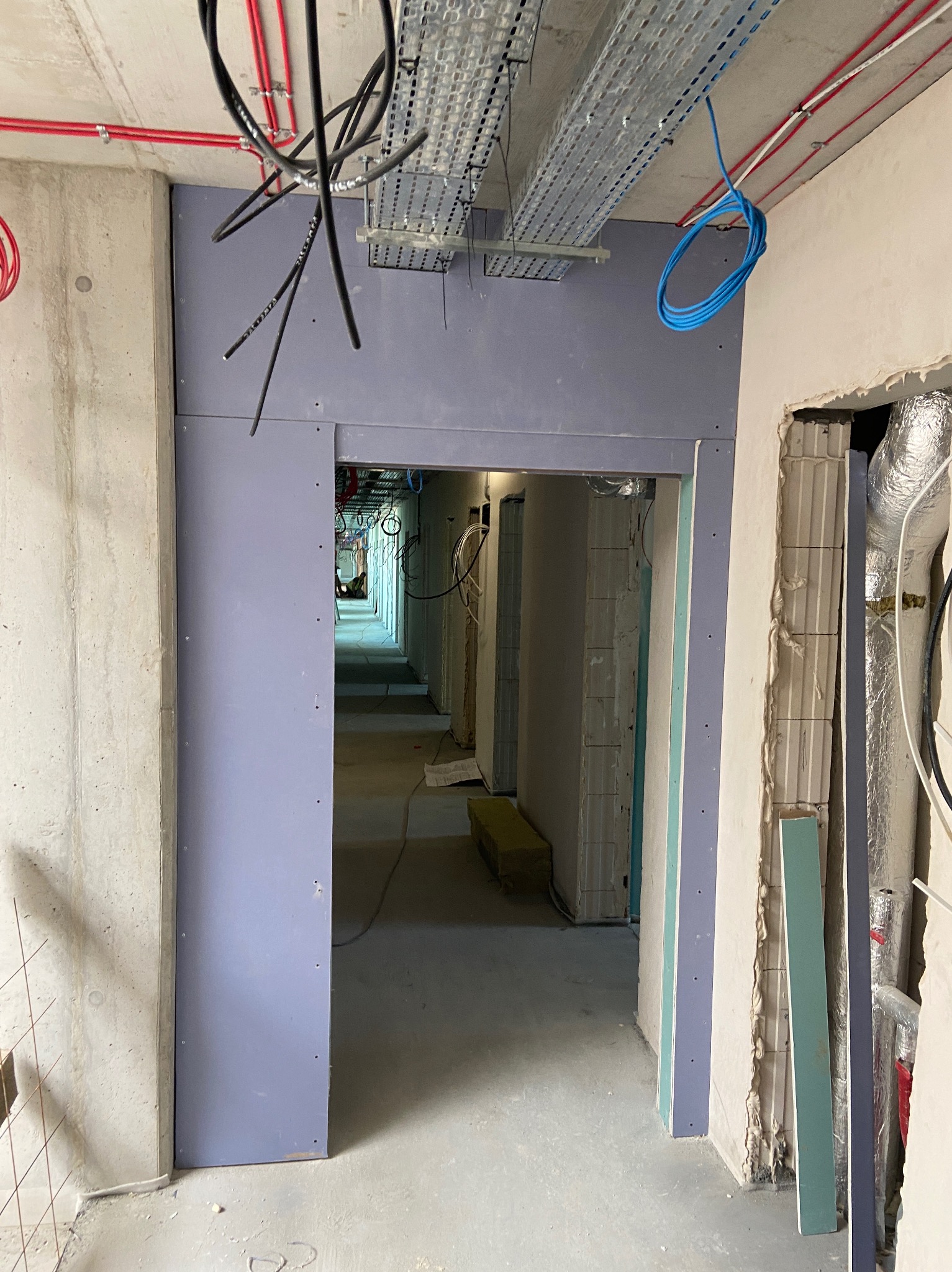 Korytarz w trakcie prac wykończeniowych: widoczne profile i płyty gipsowe, instalacje elektryczne pod sufitem. Surowy beton i elementy konstrukcyjne w tle.