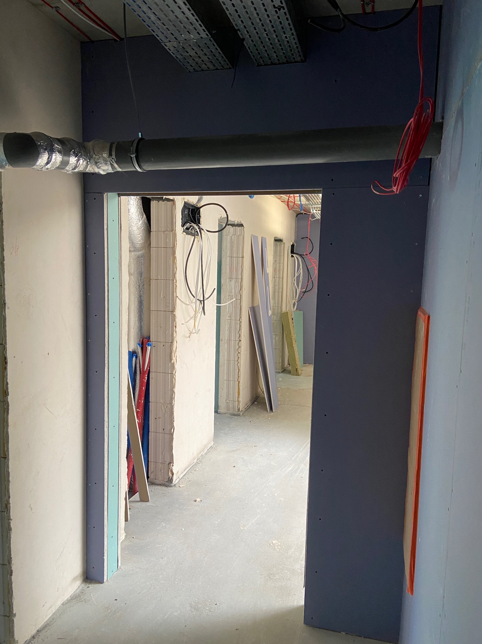 Wnętrze w trakcie prac wykończeniowych: widok przez otwór drzwiowy z zamontowaną płytą kartonowo-gipsową, widoczne instalacje i materiały budowlane w tle.