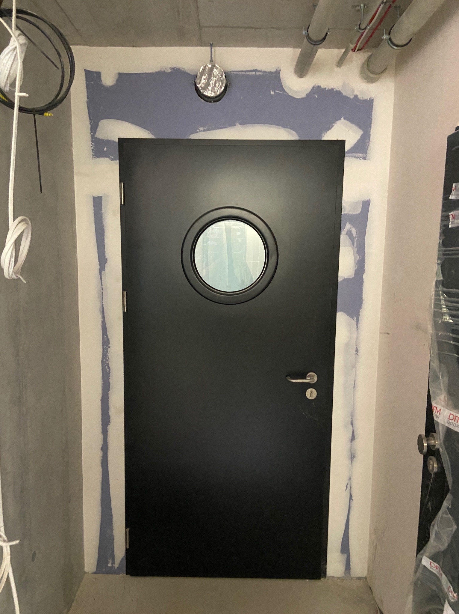 Czarna, okrągła drzwi z ościeżnicą częściowo osadzoną w ścianie, w trakcie prac wykończeniowych. Widoczne ślady szpachli i instalacje pod sufitem.