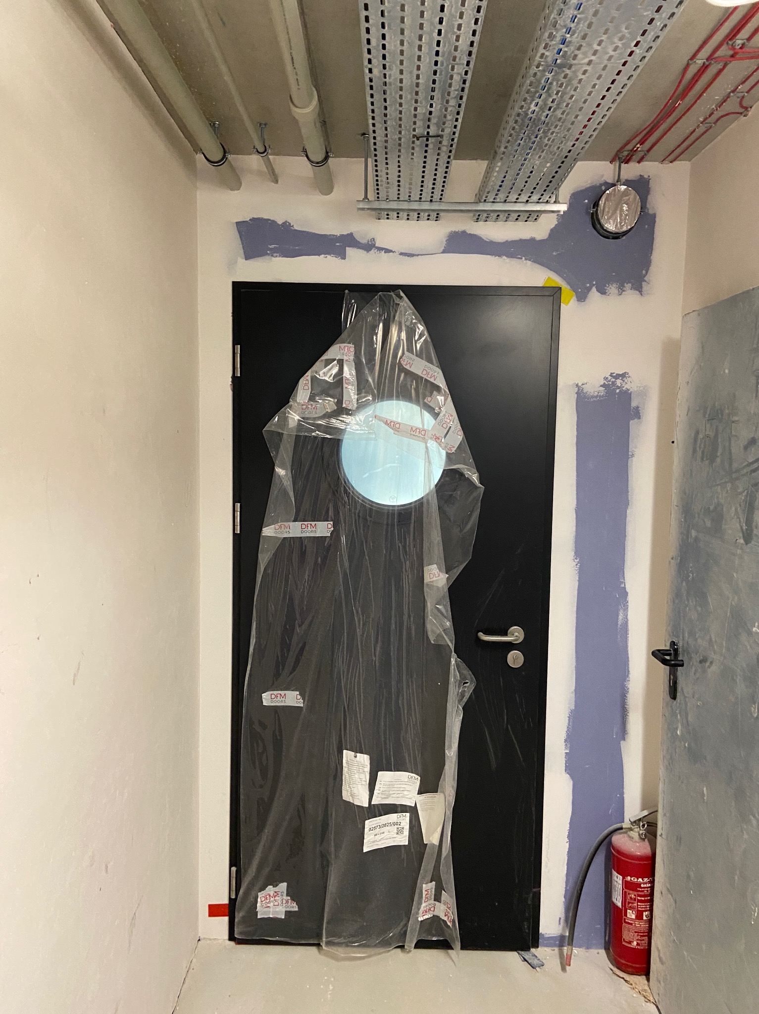 Montaż drzwi z okrągłym przeszkleniem, zabezpieczonych folią. Ściany w trakcie malowania, widoczne instalacje pod sufitem i gaśnica. Remont w lokalu użytkowym.