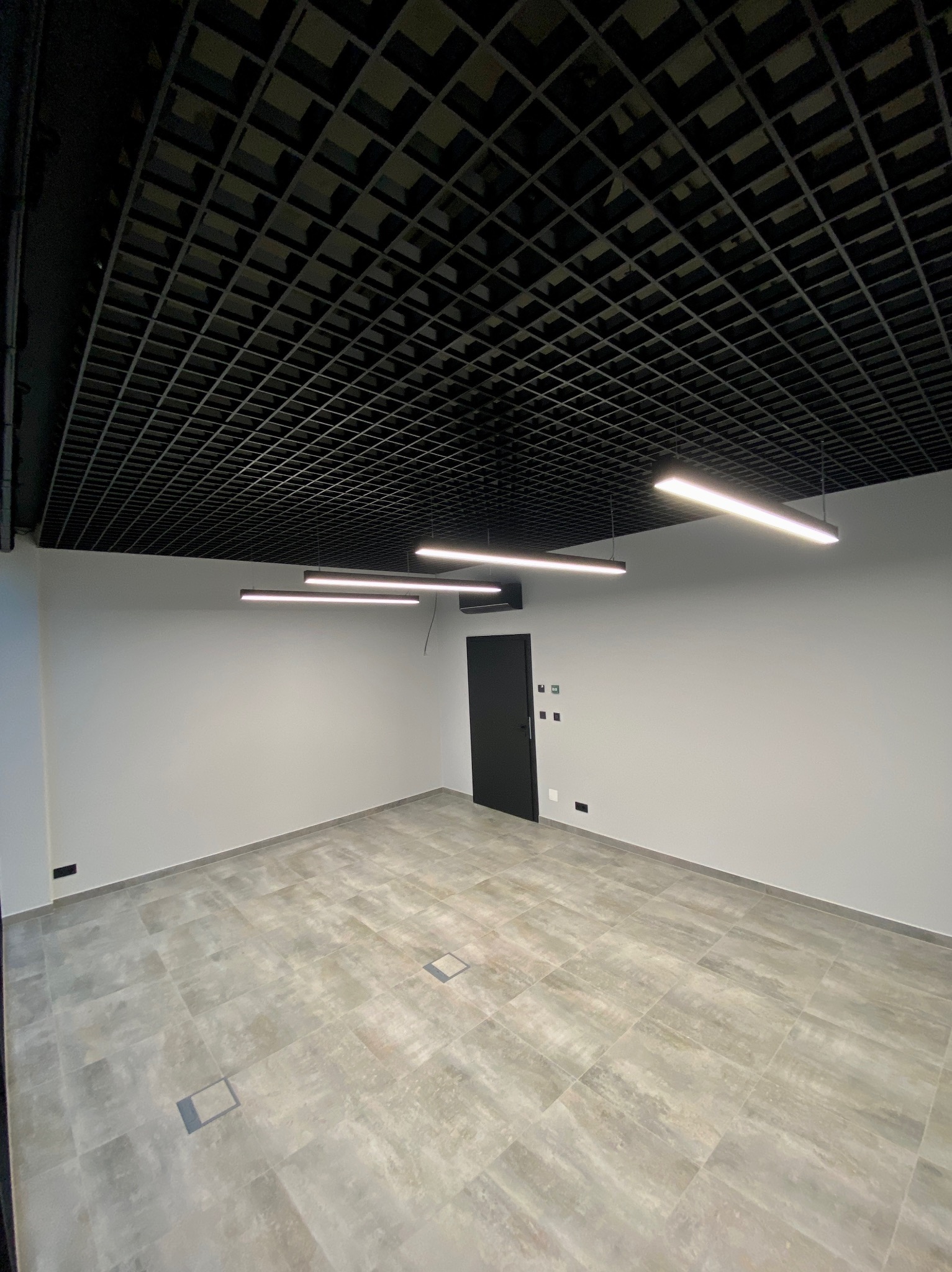 Wnętrze biurowe z podwieszanym sufitem w kratkę, szarą podłogą z płytek i minimalistycznym oświetleniem liniowym. Widoczna czarna, zamknięta  drzwi.