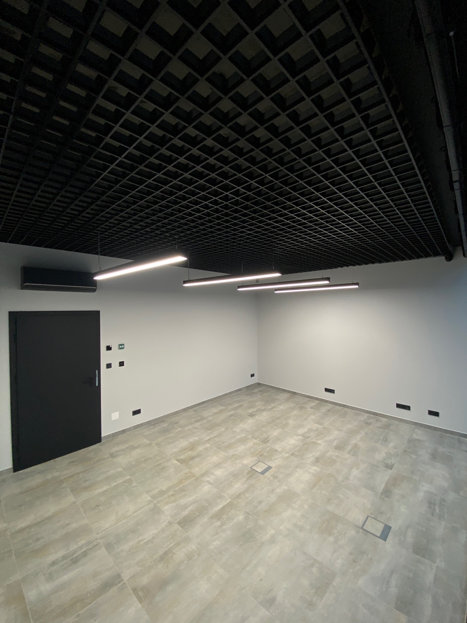 Nowoczesne wnętrze z czarną, ażurową konstrukcją sufitu, liniowymi lampami LED, szarą podłogą z płytek i minimalistycznymi ścianami. Widoczny klimatyzator i czarne drzwi.