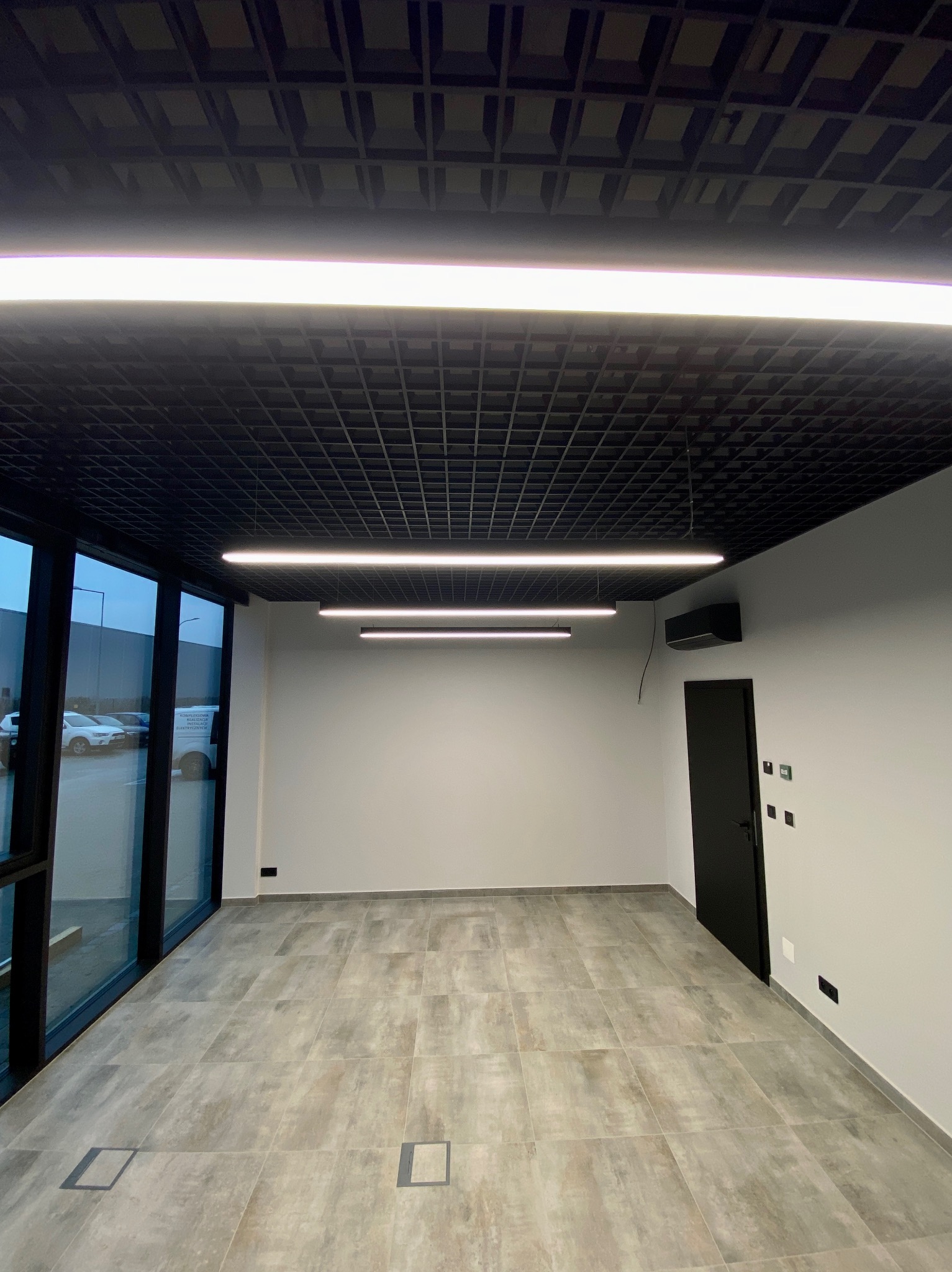 Nowoczesne wnętrze biurowe po remoncie: szara podłoga z płytek, czarny sufit rastrowy z oświetleniem liniowym LED, duże okna i czarne drzwi. Minimalistyczny design.