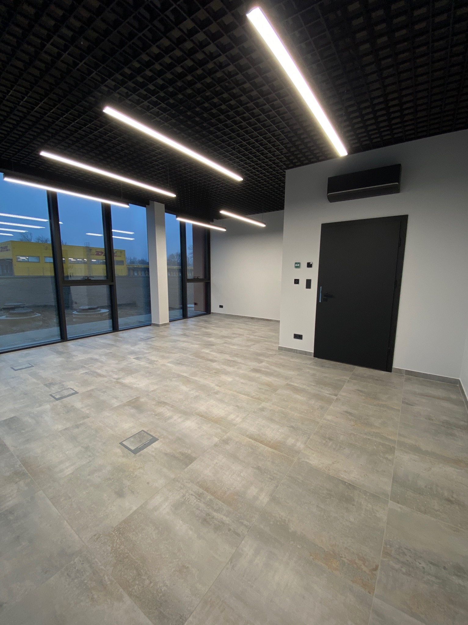 Nowoczesne, przestronne wnętrze biurowe z szarą podłogą z płytek, czarnymi drzwiami i oknami z widokiem na zewnątrz. Oświetlenie sufitowe w postaci długich, białych lamp.