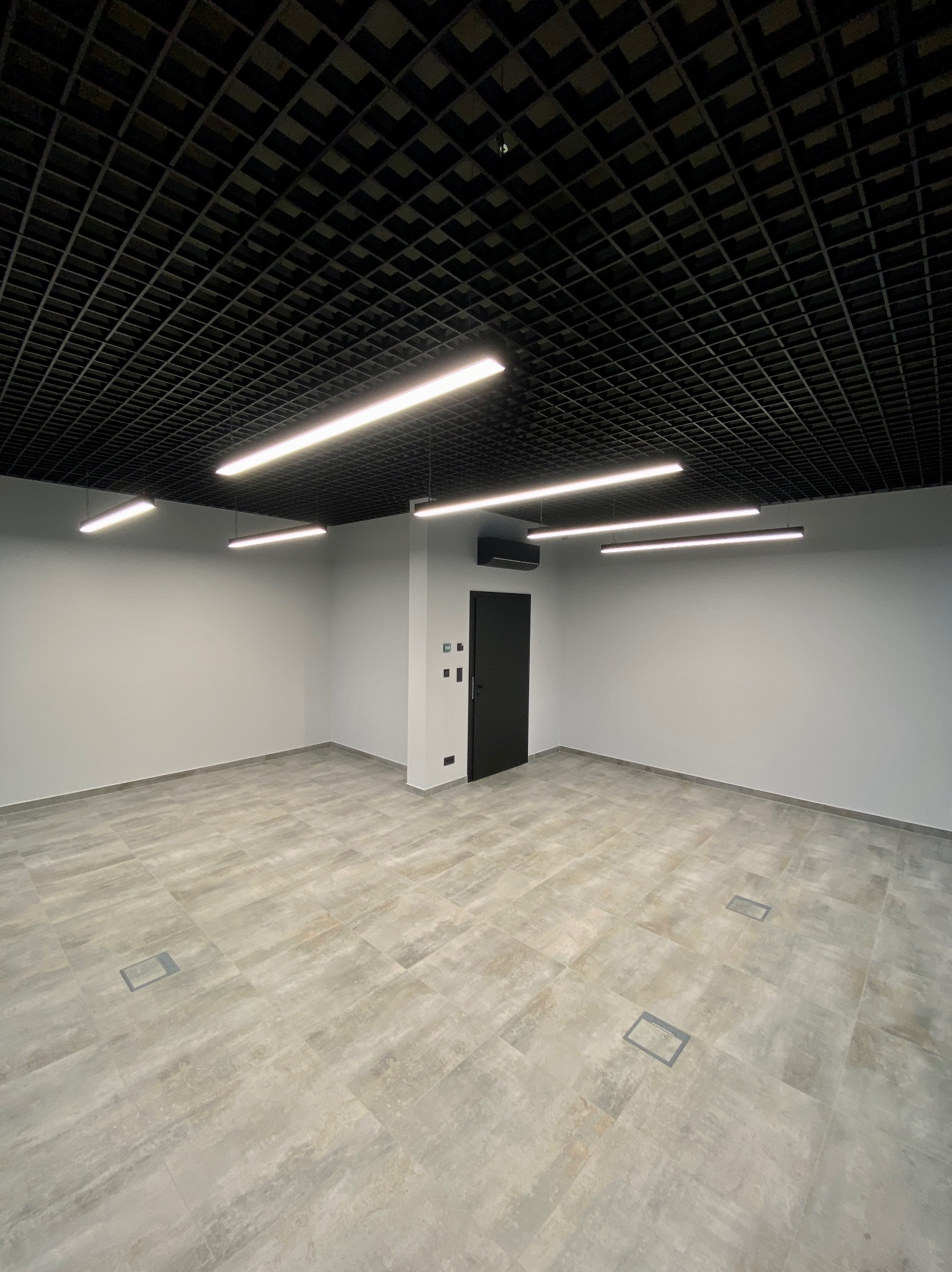 Jasne płytki podłogowe w przestronnym, nowocześnie wykończonym wnętrzu z czarnym sufitem rastrowym i liniowym oświetleniem LED. Widoczna czarna ściana z drzwiami.